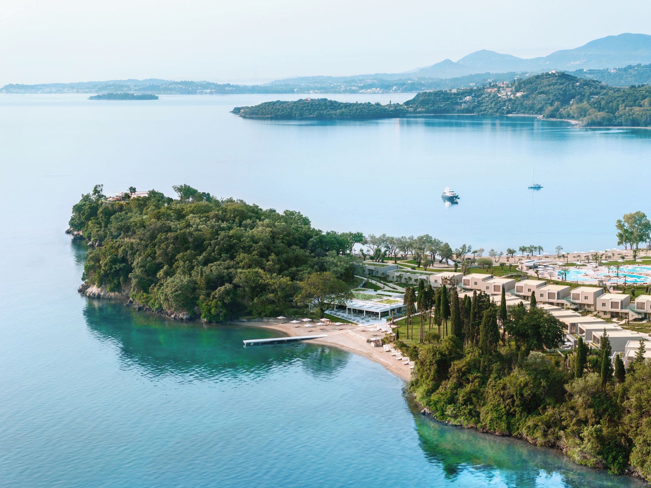 Aerial view of a waterfront residential area with trees, modern houses, and boats on calm water, with mountains in the background.