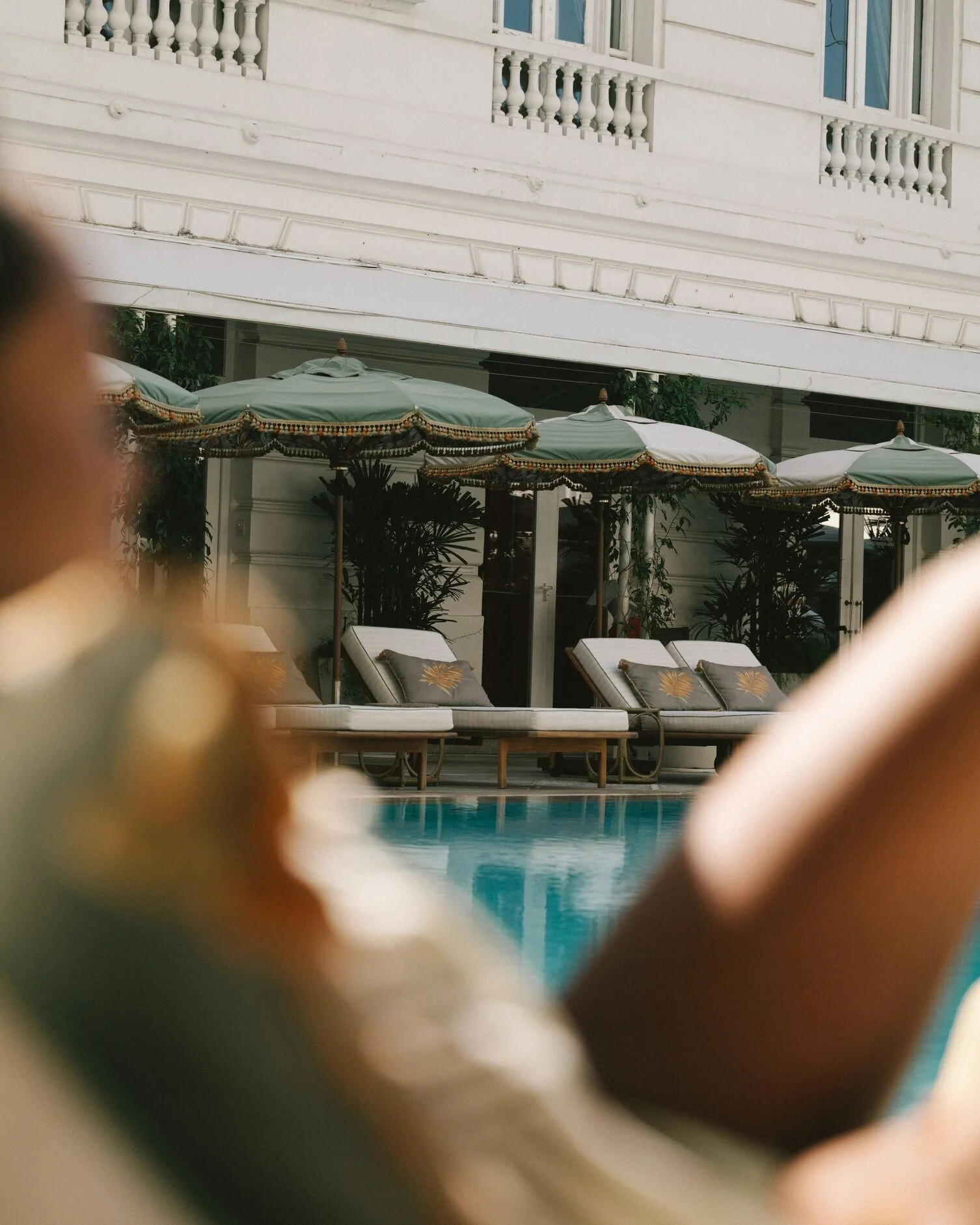 View of poolside lounge chairs with pillows, surrounded by umbrellas, in front of a white building with balconies.