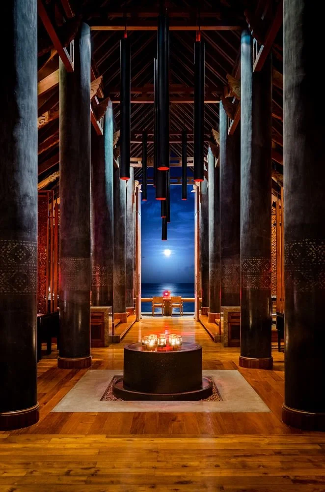 Interior of a spacious, wooden hall with tall black columns, pendant lights, and a view of the moon over the ocean in the background. Candles are placed on a round table on a rug in the foreground.