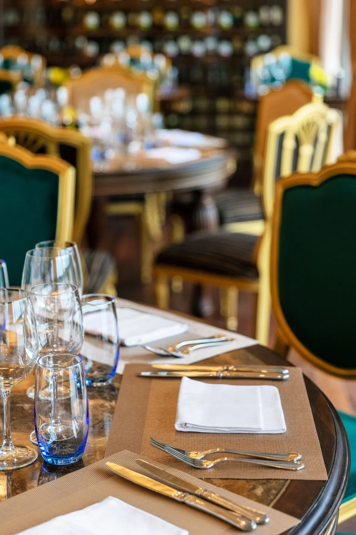 A formal dining table set with white napkins, silverware, and empty wine glasses, in a restaurant with green chairs and a bookshelf in the background.