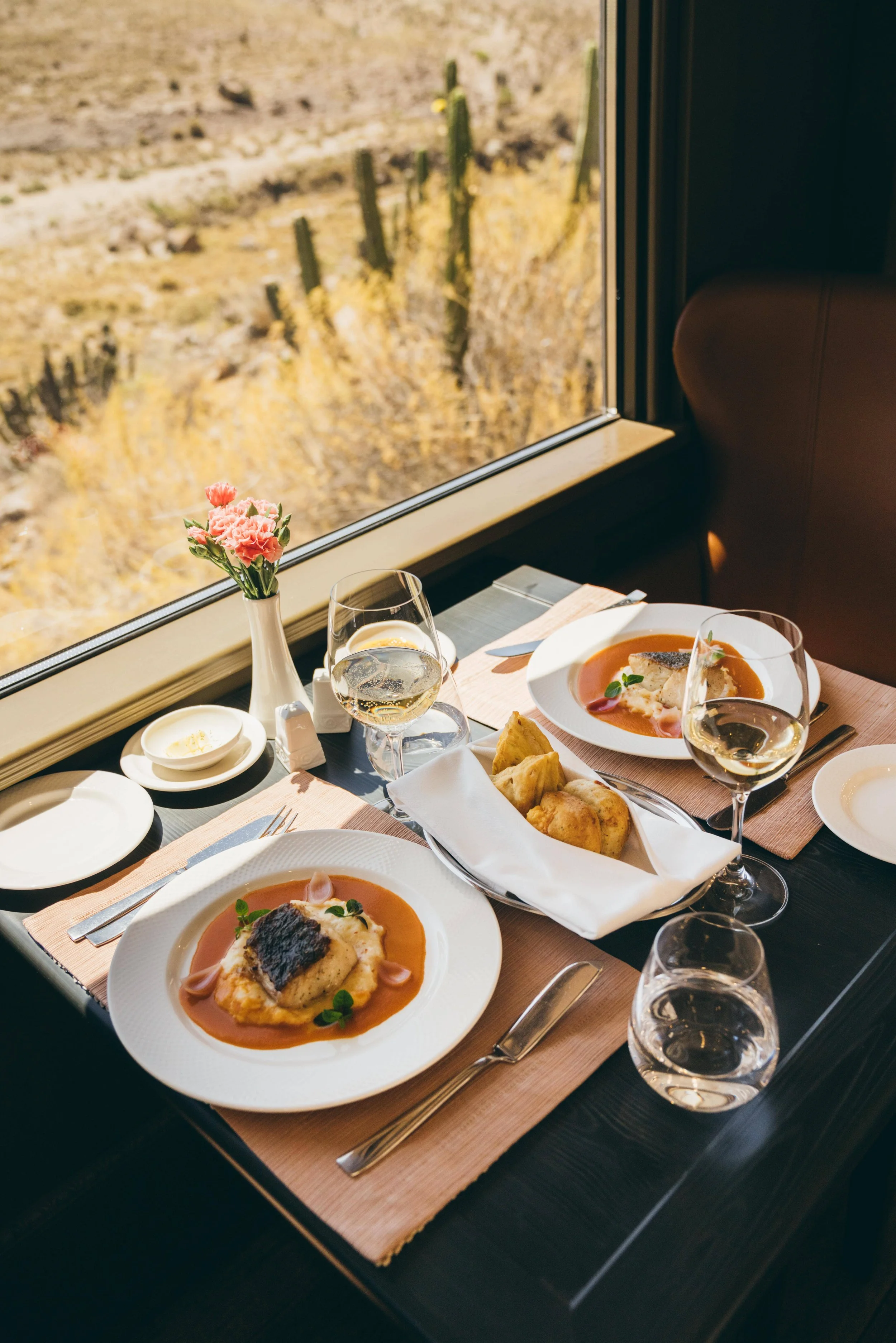 A table set for dining with plates of food, glasses of white wine, and a small vase of pink carnations. The scene is near a large window with a desert landscape featuring cacti visible outside.