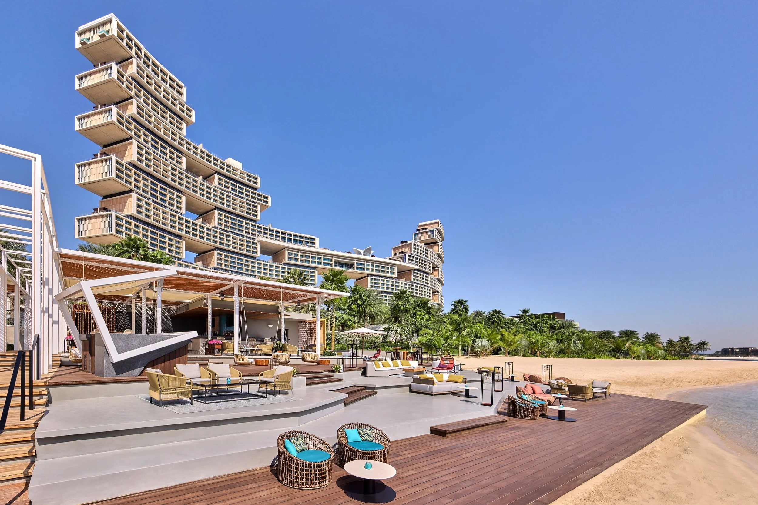 Luxury hotel with unique stacked balconies, outdoor lounge area with chairs, tables, and umbrellas, beachfront with sand and jungle of palm trees, clear blue sky.