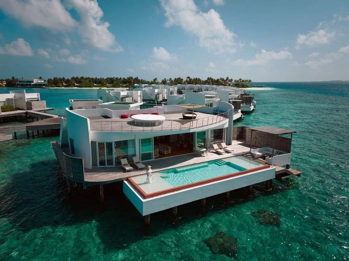 Overwater villa with private pool and deck, situated on clear blue ocean, with modern white architecture, patio lounge chairs, and a person standing near the pool.