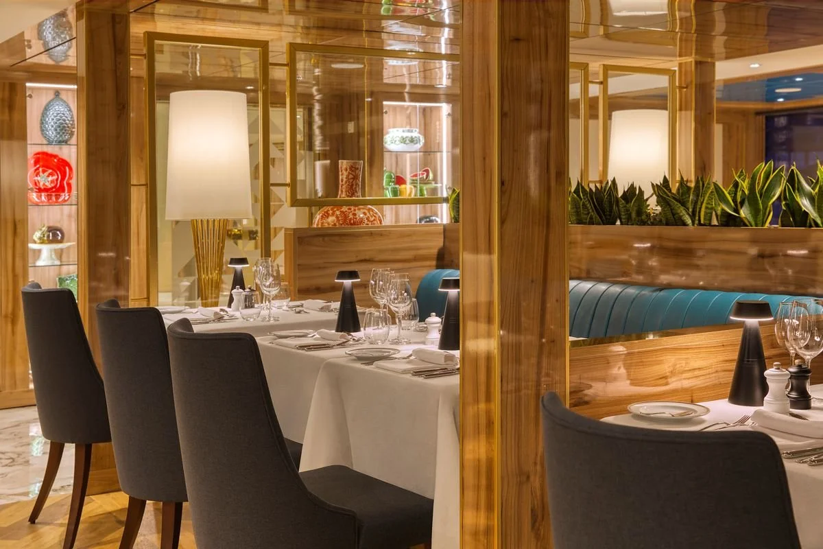 A restaurant with a long table covered in a white tablecloth, set with glassware and tableware, surrounded by black chairs. Wooden partitions and shelves with decorative bowls and plants enclose the dining area. Soft lighting creates a warm ambiance.