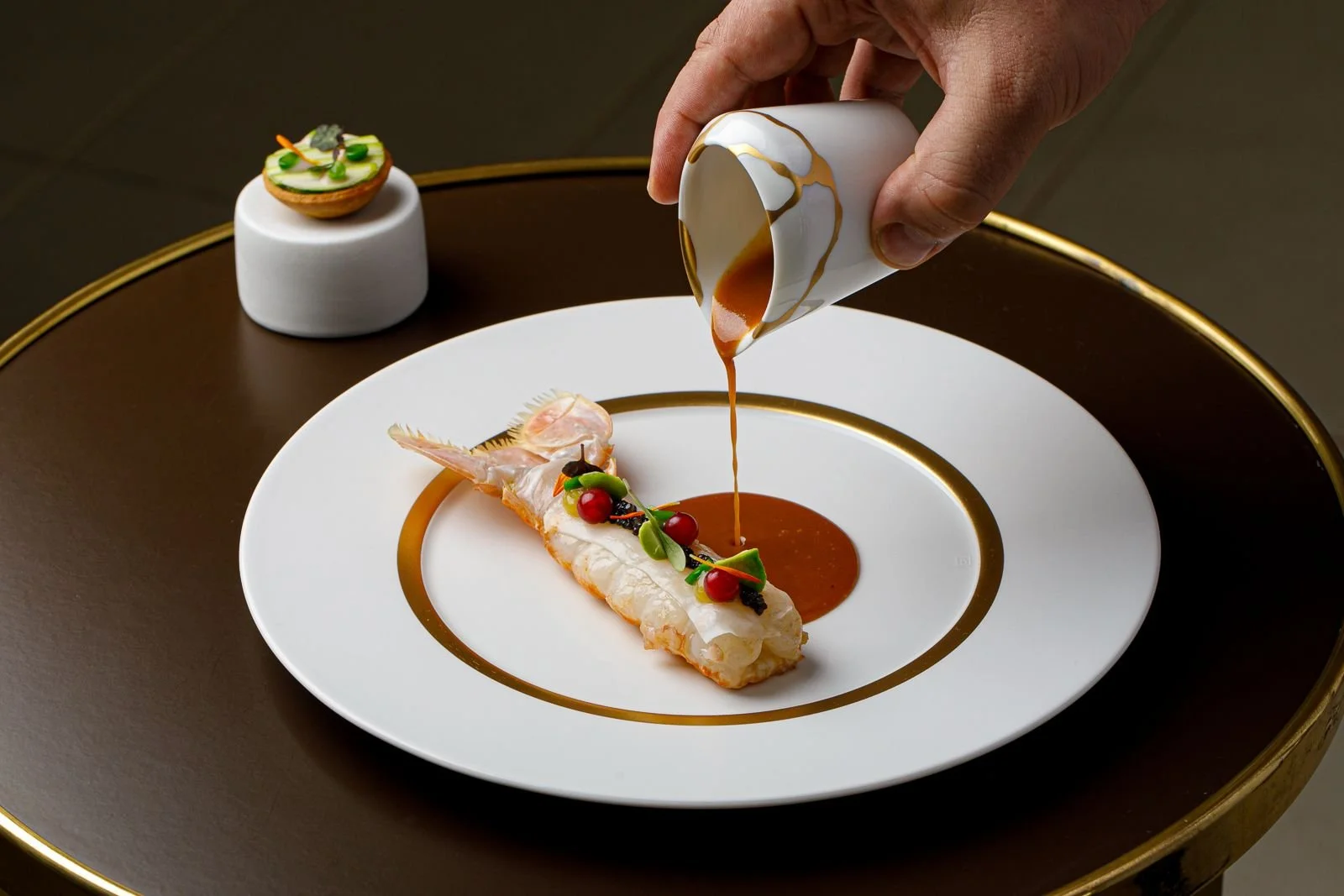 A gourmet dish featuring a cooked shrimp garnished with small red berries and greens on a white plate with gold accents. A hand is pouring sauce from a small white cup over the shrimp, with a small dish of decorated appetizer in the background.