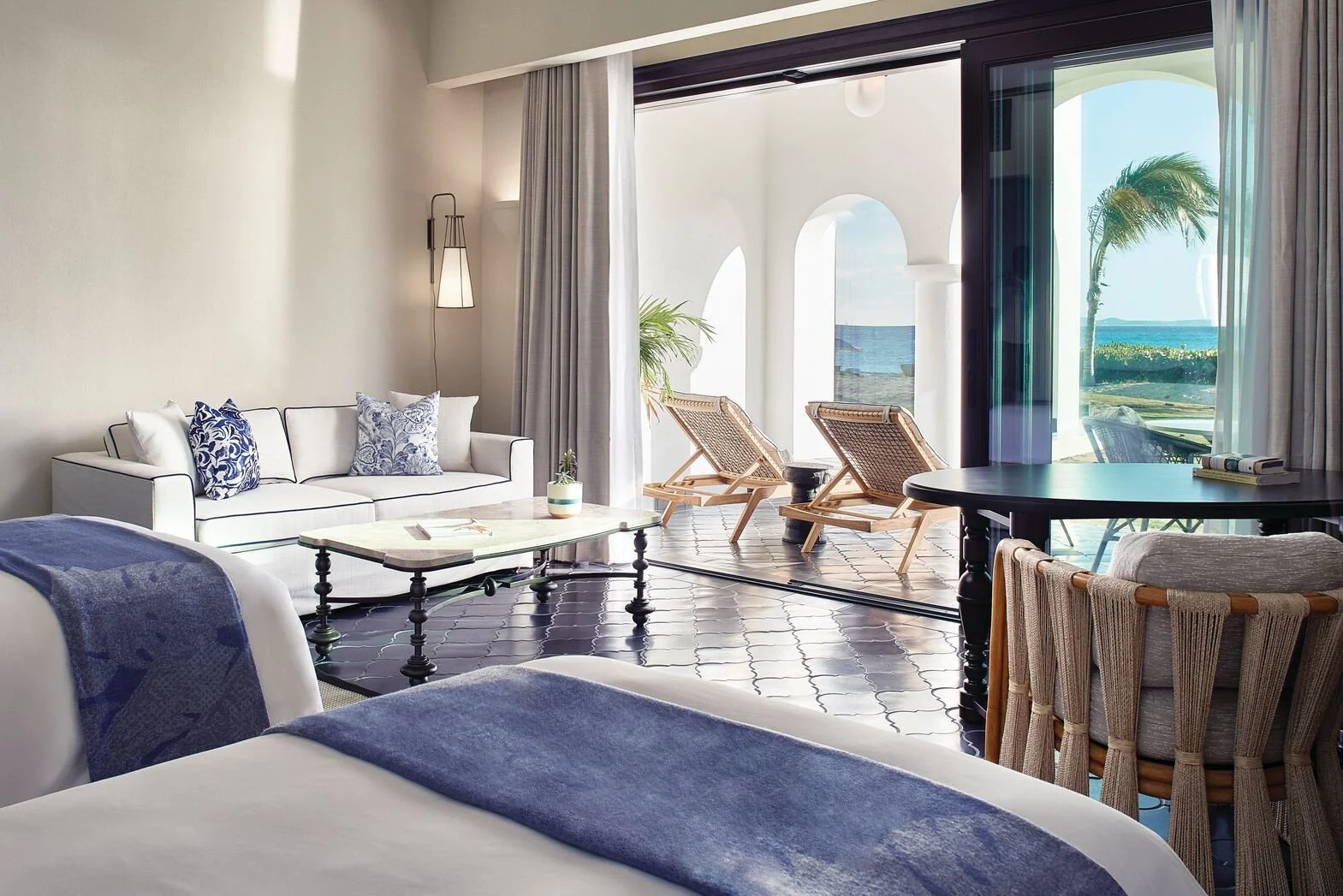 Living room with white sofa, blue pillows, coffee table, and a balcony with lounge chairs overlooking the ocean and palm trees.