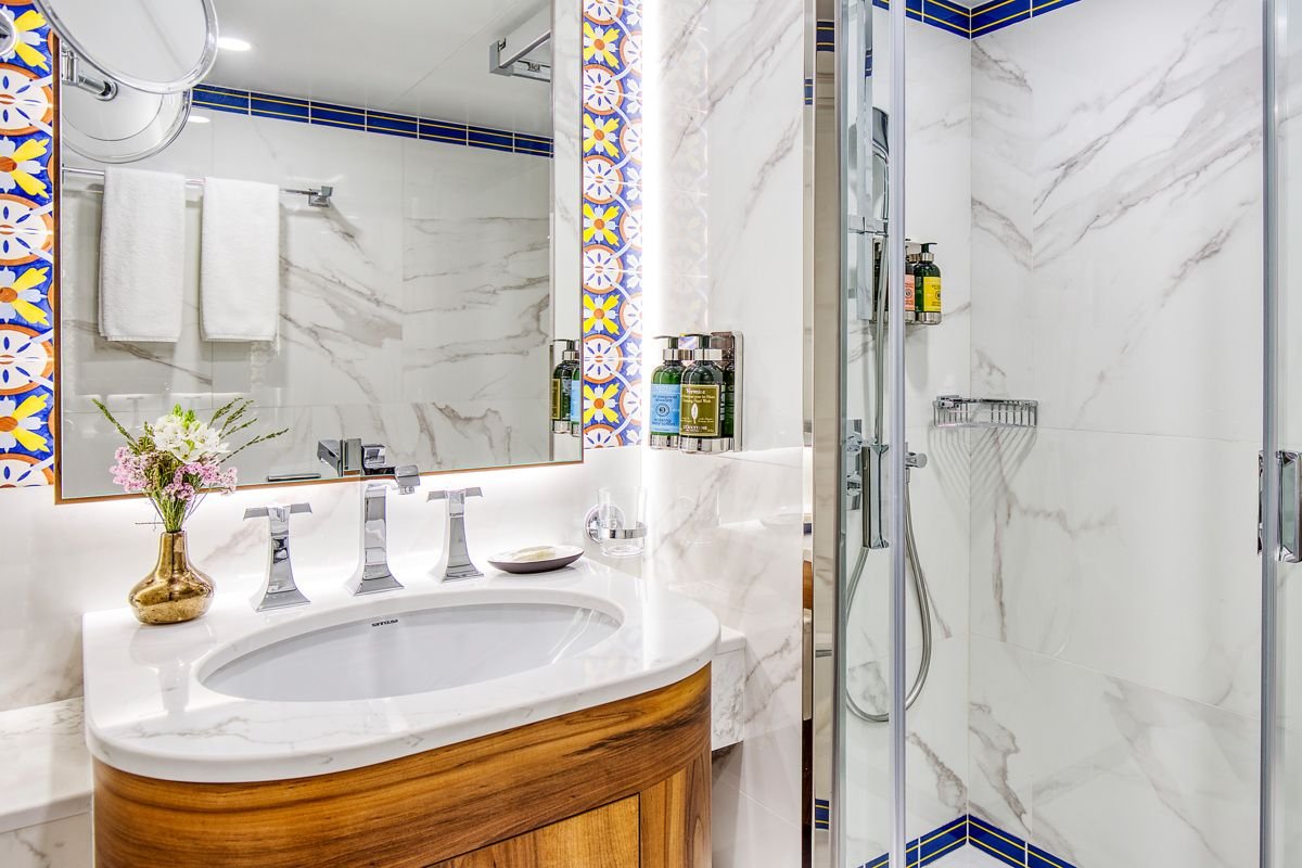 A modern bathroom with a marble sink countertop, a large mirror with decorative blue, yellow, and white tiles framing it, a glass shower stall, and some toiletries and towels.