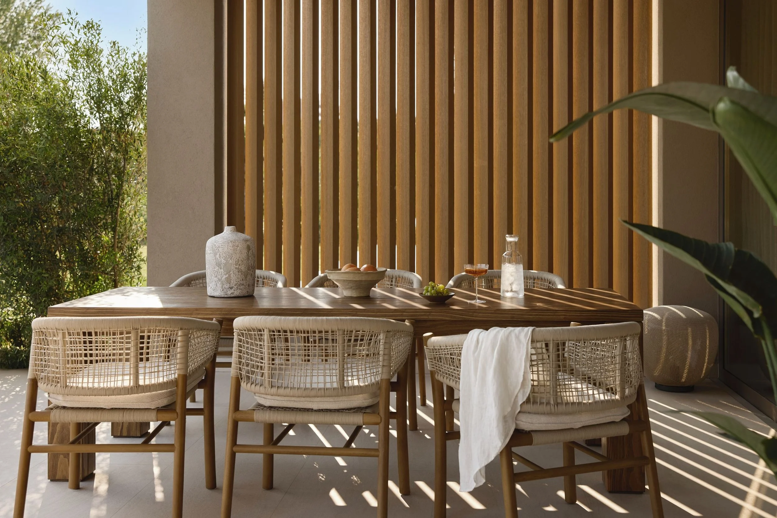 Outdoor dining area with wooden slat wall, wicker chairs, and a wooden table with a ceramic vase, a fruit bowl, a glass bottle, and a glass of drink, with sunlight creating shadows.