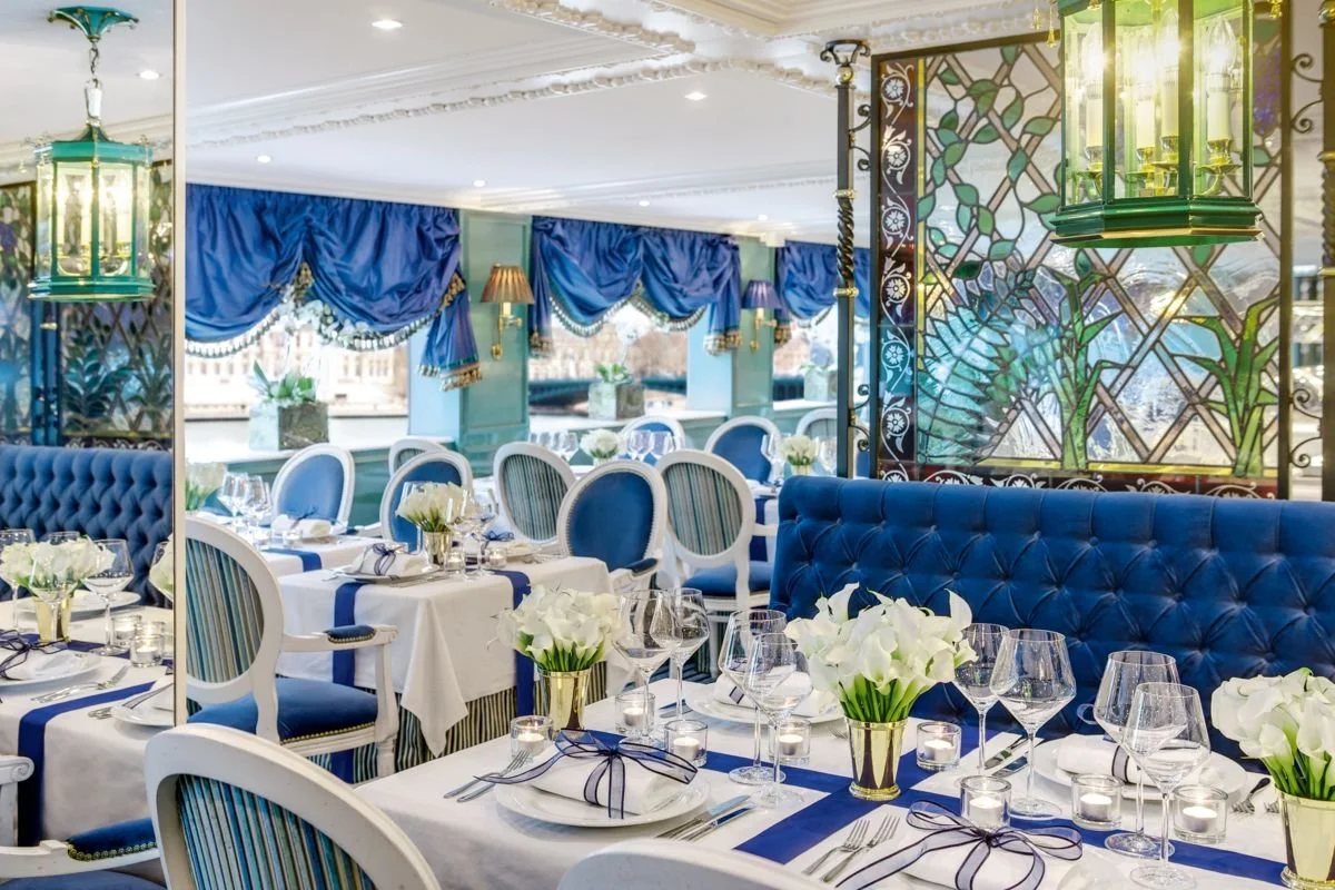 Elegant dining room with white tablecloths, blue and white striped chairs, floral centerpieces, and blue drapery, with decorative stained glass window and green chandeliers.