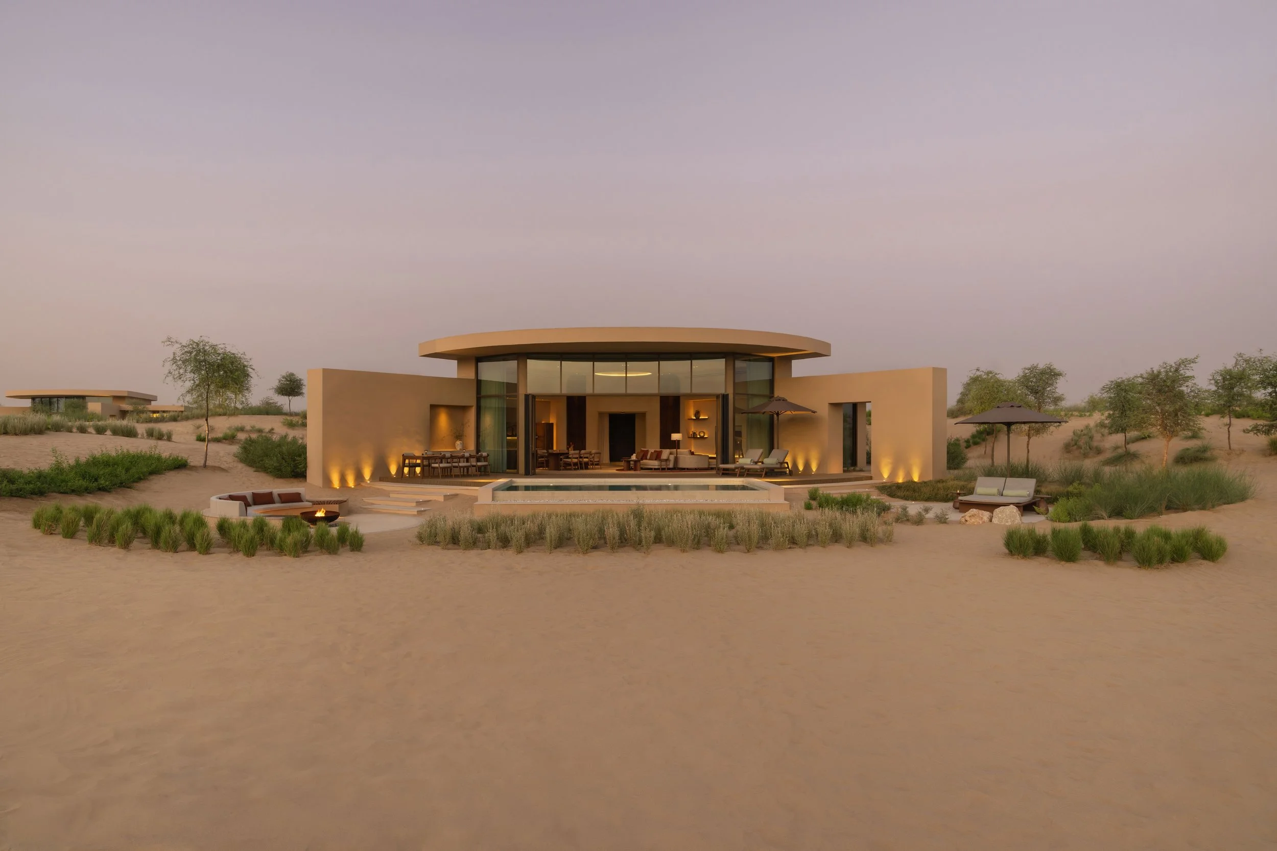 Modern house with curved roof, large glass windows, outdoor furniture, in a desert landscape at sunset with sand and sparse trees.