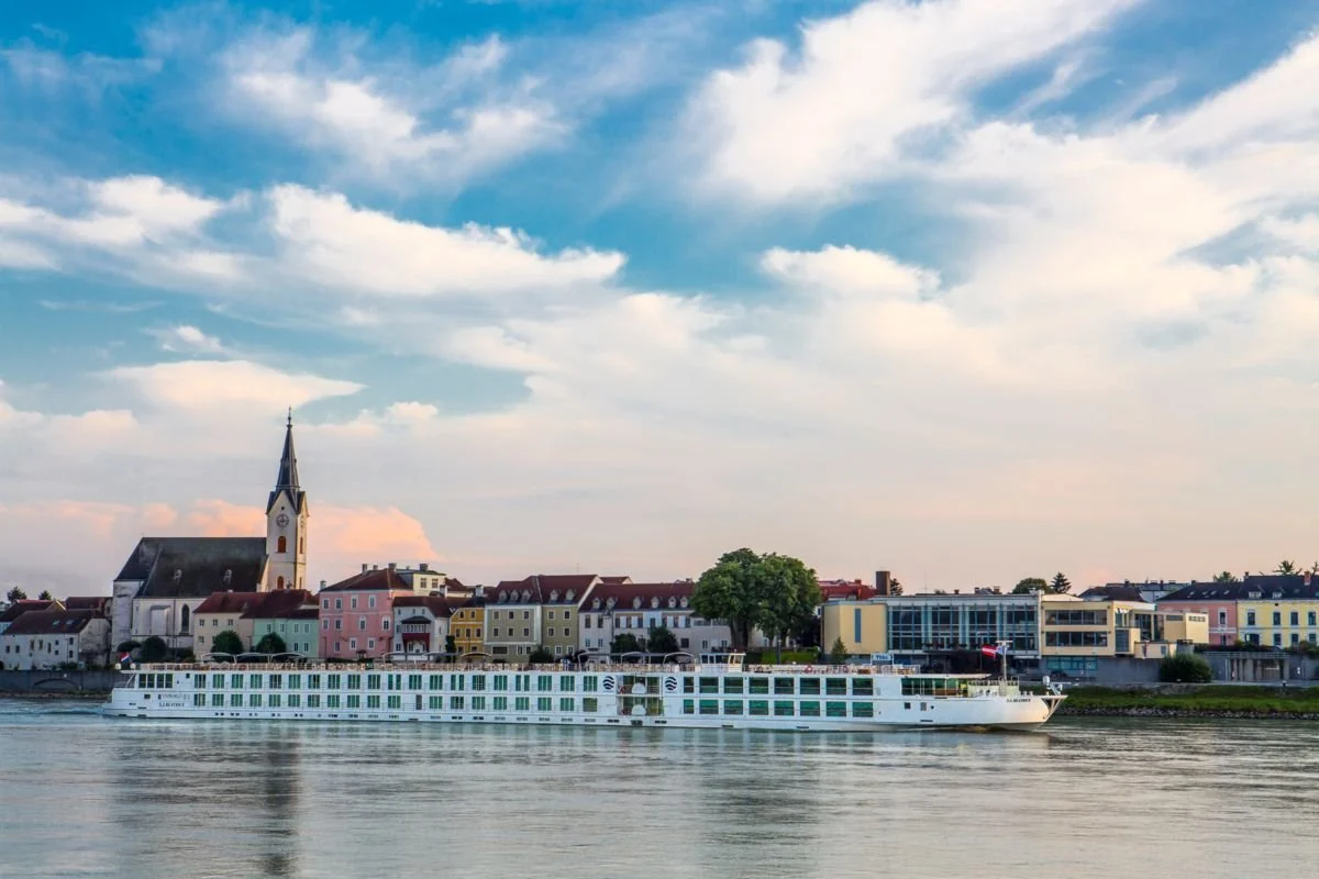 A river with a large white cruise ship passing by a town with colorful buildings and a church with a tall steeple, under a sky with scattered clouds.