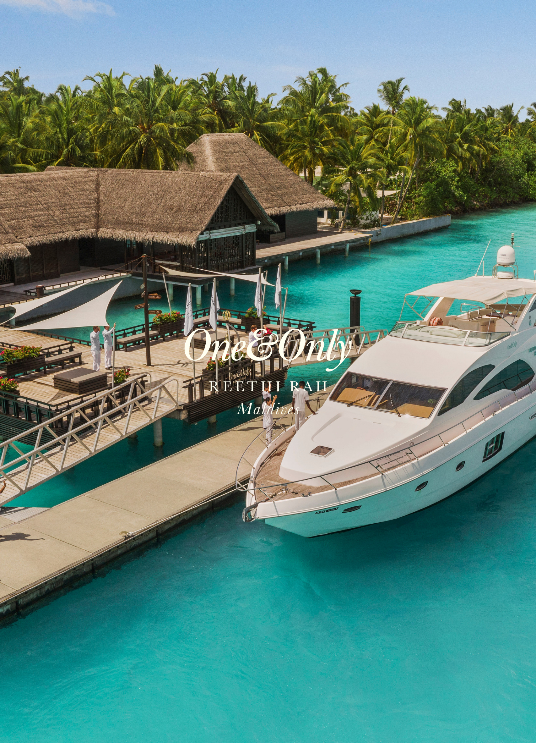 A luxury yacht docked at an overwater resort in the Maldives, with a thatched-roof bungalow, palm trees, and clear turquoise water.