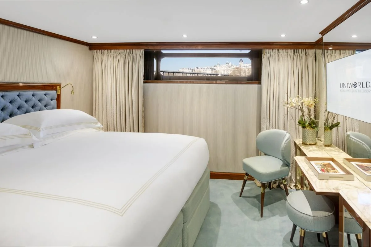 Hotel room with a bed, curtains, a window view, a small table with chairs, and a wall-mounted TV.
