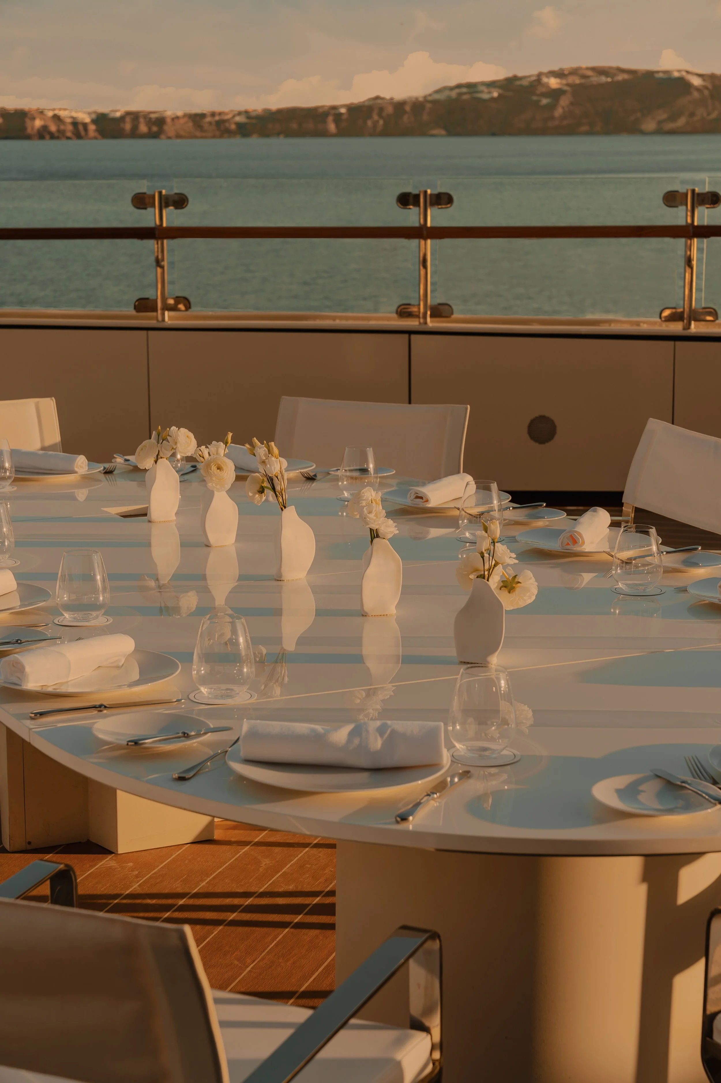 An outdoor dining area on a boat with a view of the ocean and cliffs in the distance; the table is set with white plates, napkins, glasses, silverware, and small white vases with white flowers, during sunset.