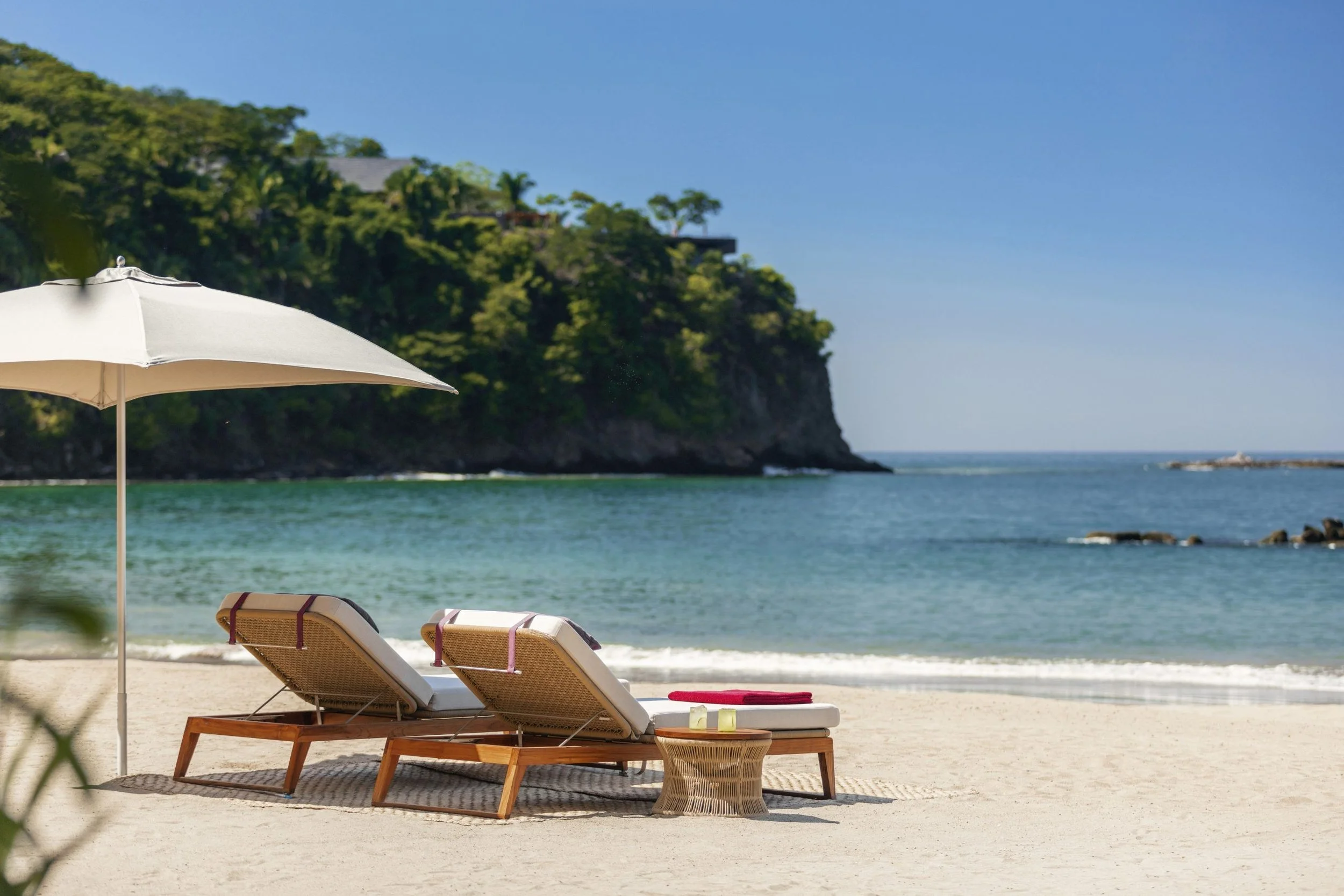 Beach scene with two lounge chairs under an umbrella, facing the ocean with a rocky shoreline and a green cliff in the background under a clear blue sky.