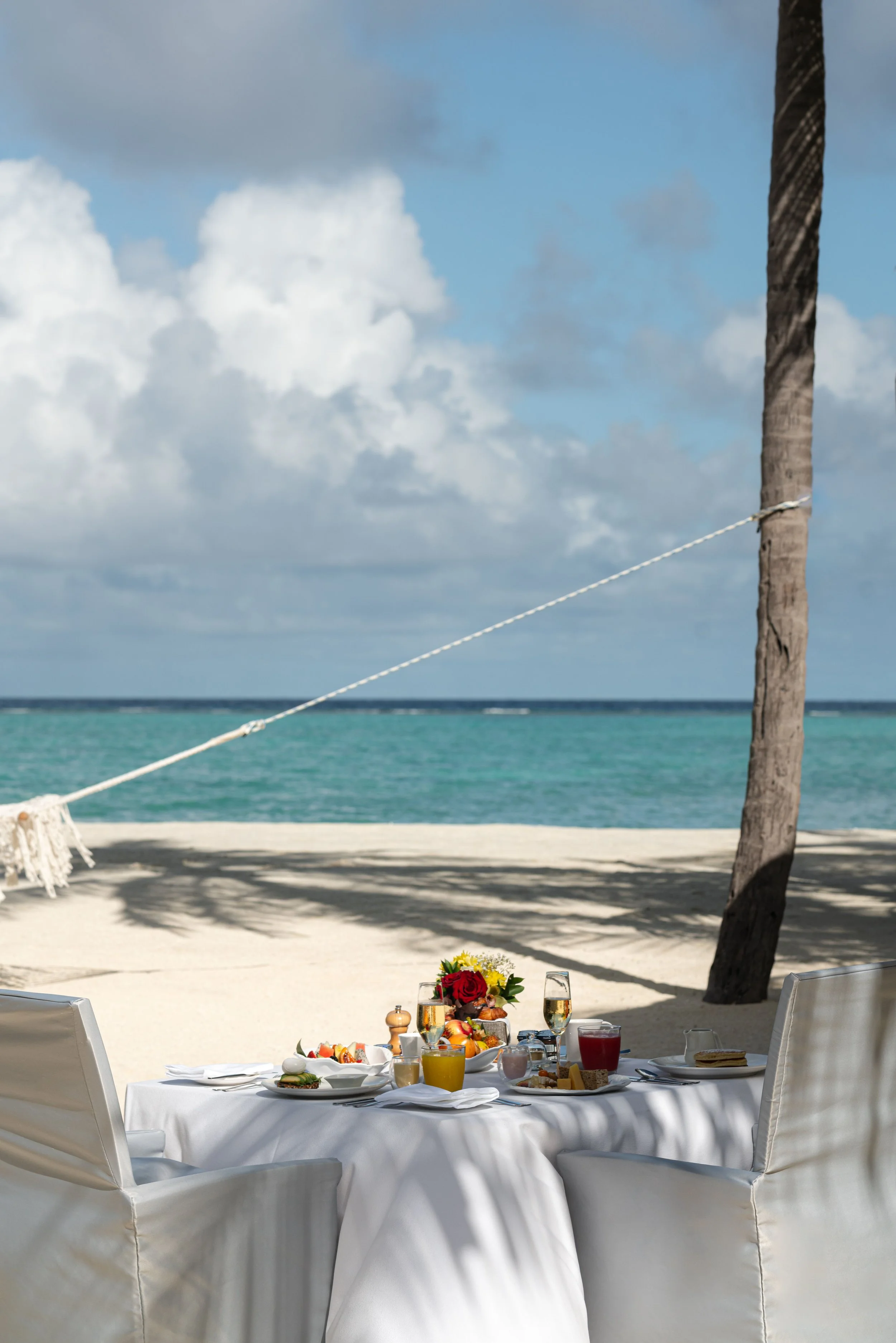 A beachside table set with breakfast foods, drinks, and flowers, with the ocean and sky in the background.