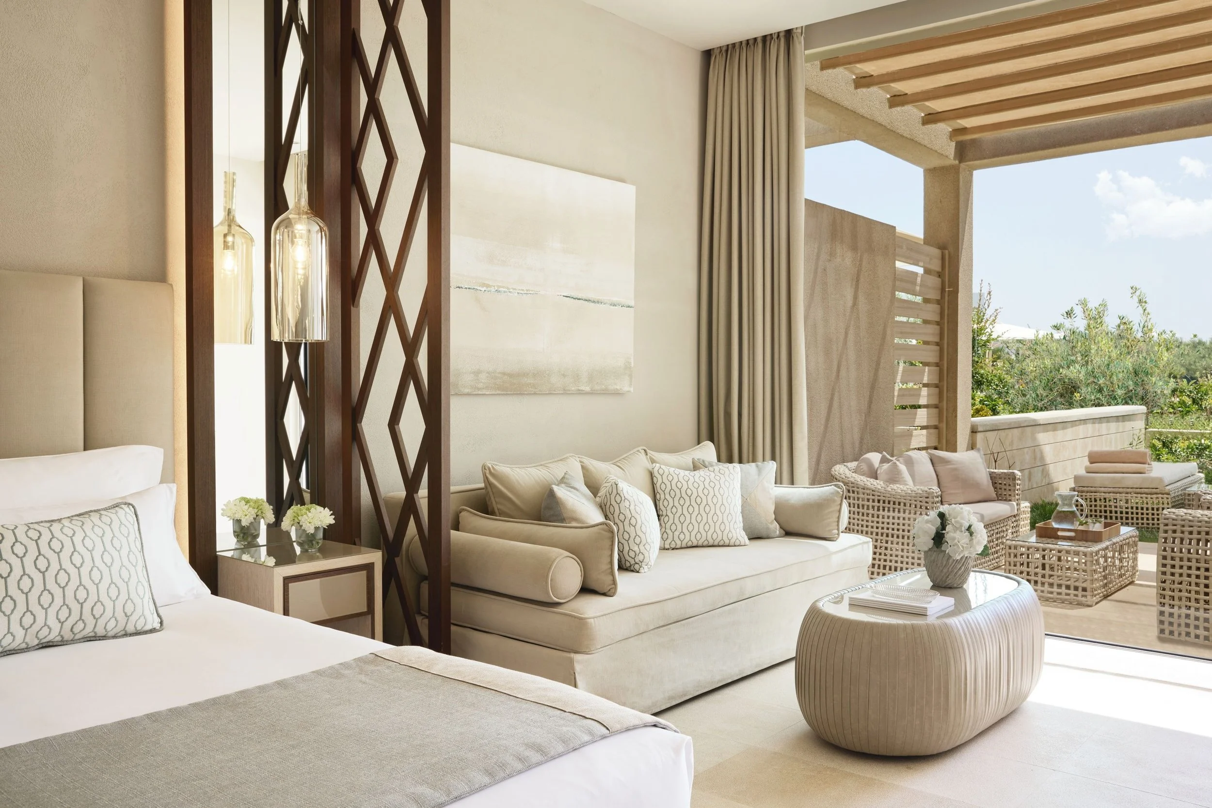 Bright, modern living room with white and beige furniture, patterned cushions, glass pendant lighting, artwork on wall, and an outdoor patio with seating and greenery visible through large open sliding doors.