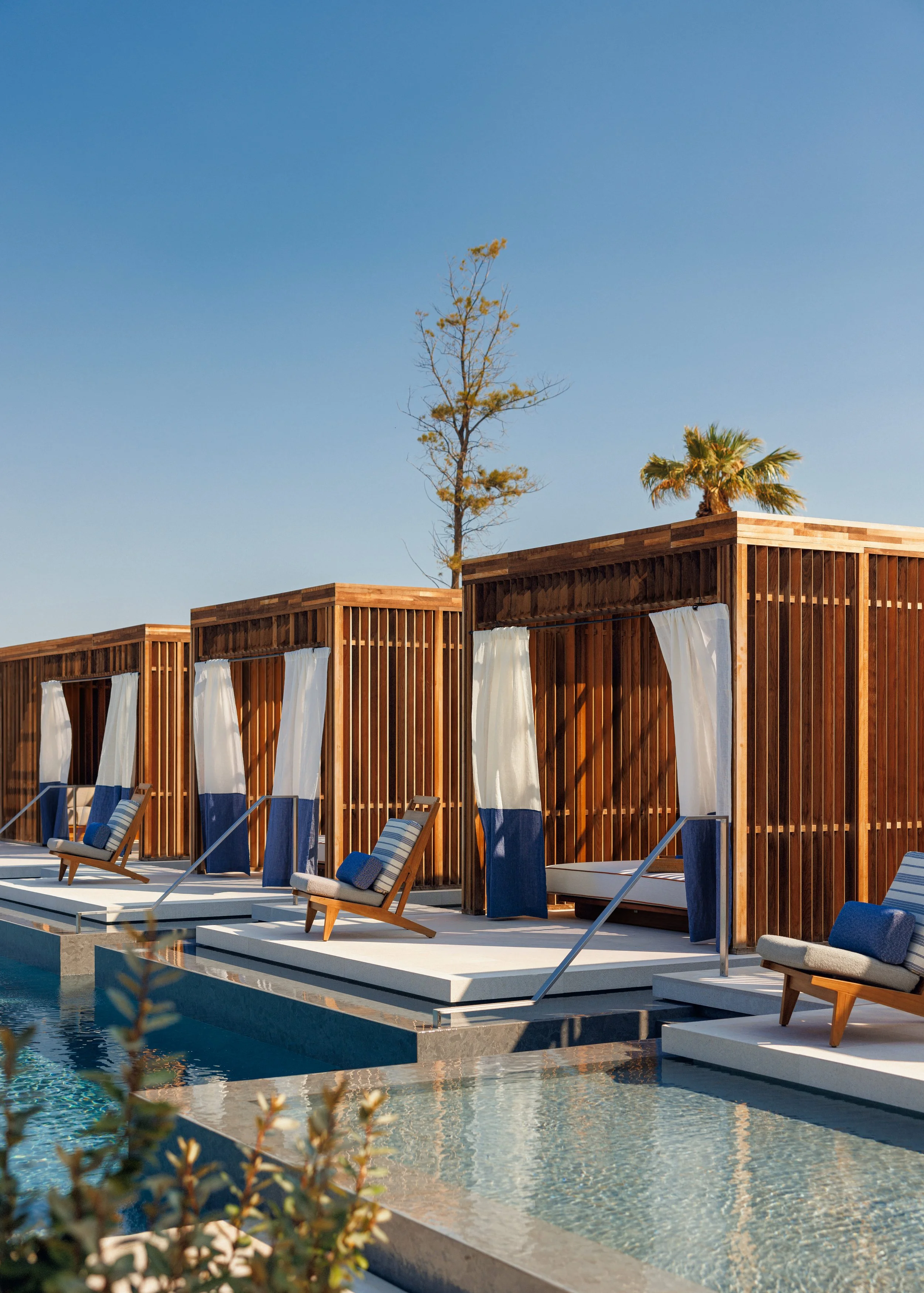 Poolside cabanas with wooden slats and white curtains, blue and beige lounge chairs, overlooking a swimming pool with clear water, two tall trees in the background, under a clear blue sky.