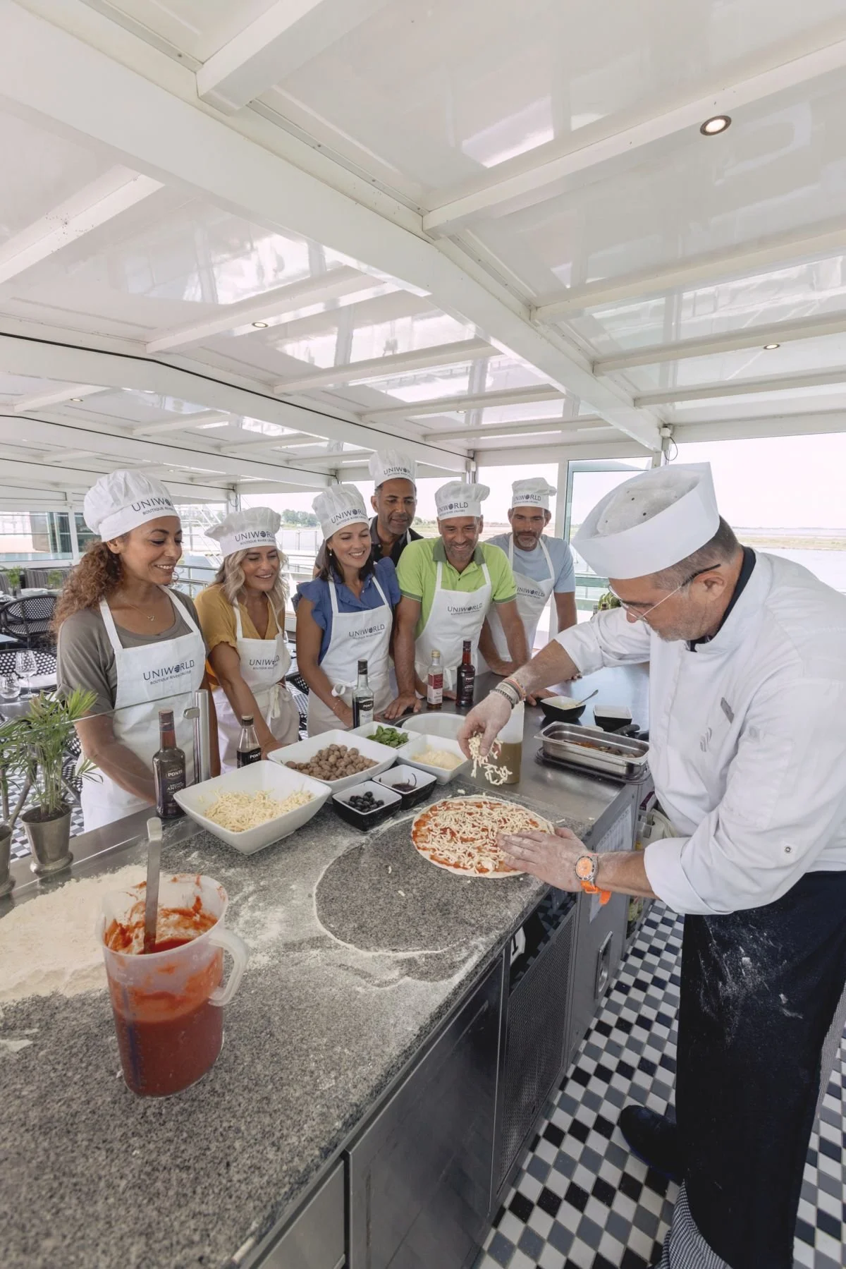 A group of six people in chef hats and aprons standing around a counter watching a chef prepare a pizza. The counter is filled with bowls of ingredients like cheese, meat, and vegetables, with a pizza in progress.