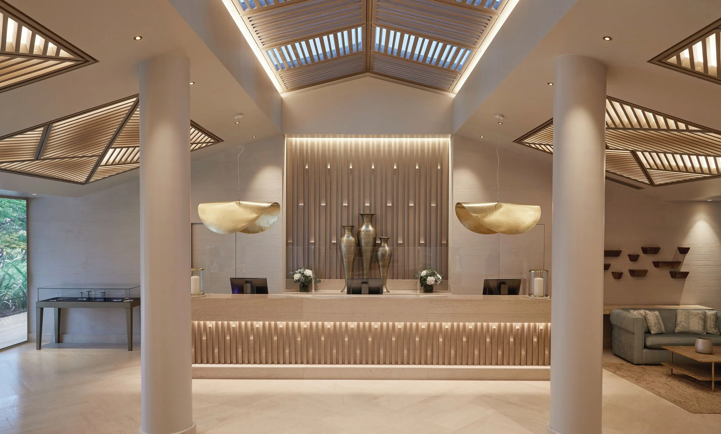 Modern hotel lobby with a reception desk, decorative vases, gold-colored pendant lights, and horizontal wooden slats on the wall with soft lighting. There are large white columns and a ceiling with geometric wooden panels and recessed lighting.