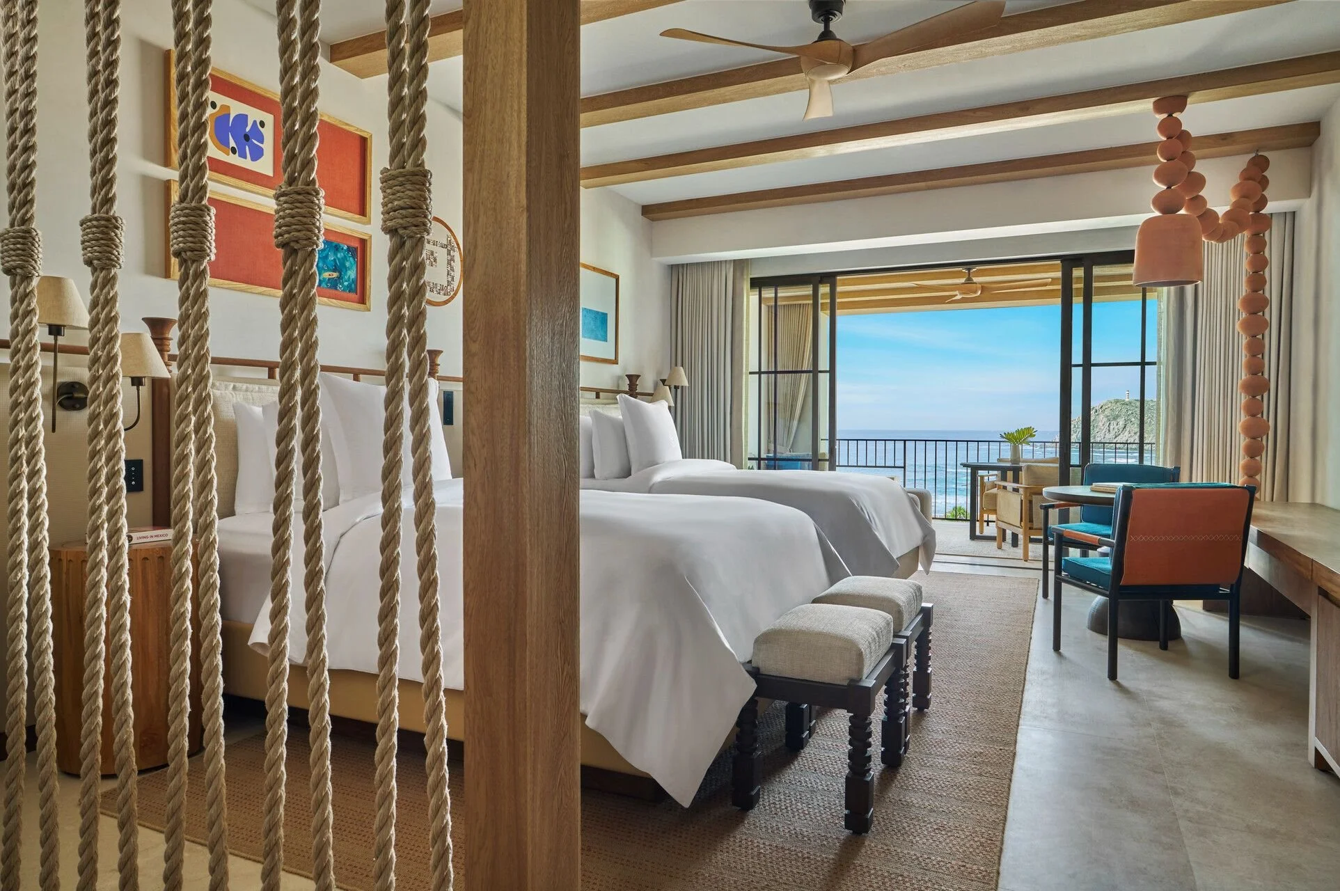 A hotel room with two beds, artwork on the wall, and large windows showing a view of the ocean and a rocky hill.