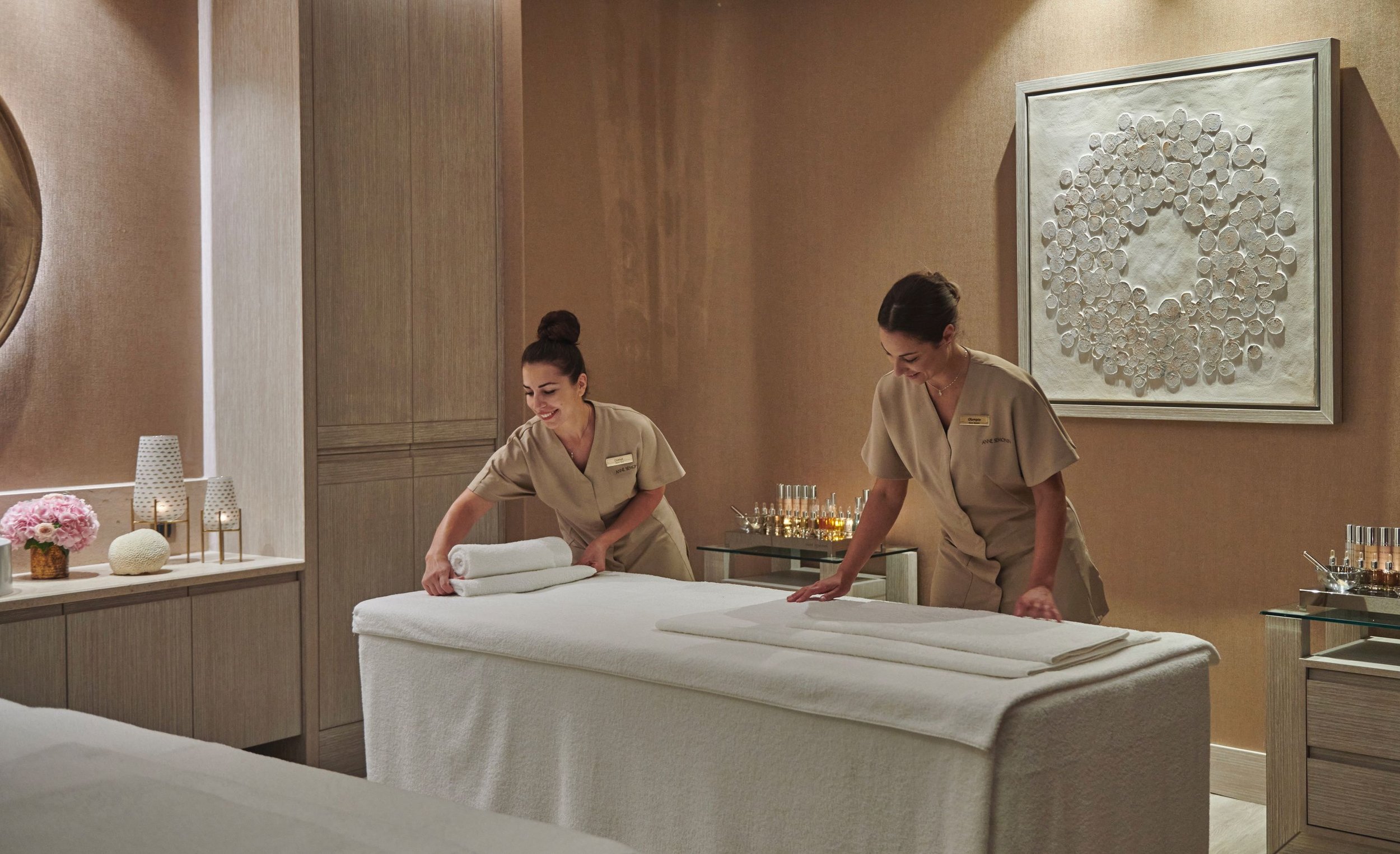 Two women in spa uniforms preparing a massage table with towels in a calming spa room with decorative art and flowers.