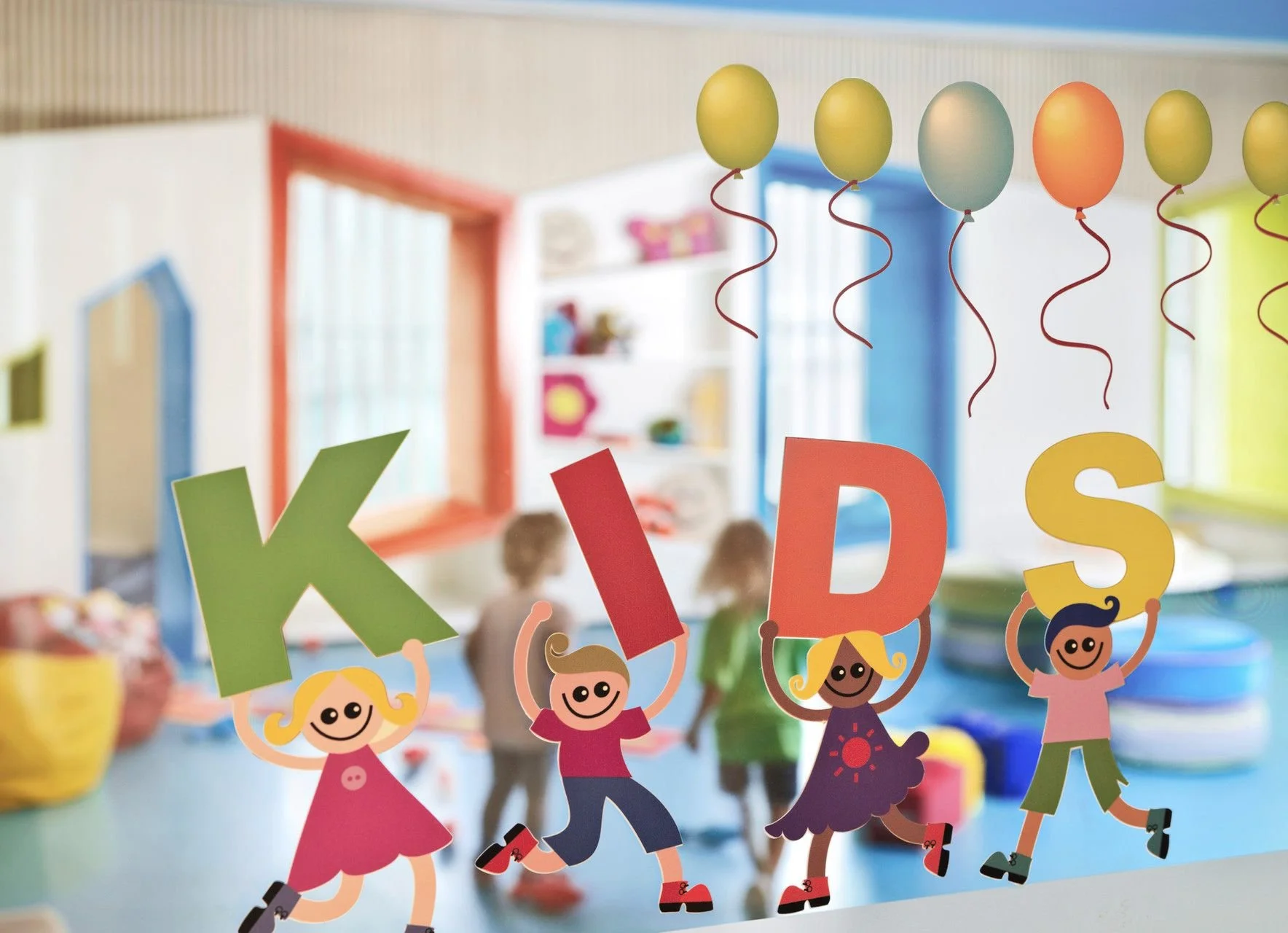 Colorful paper cutouts of children holding letters spelling 'KIDS' on a classroom window, with balloons and children playing in the background.