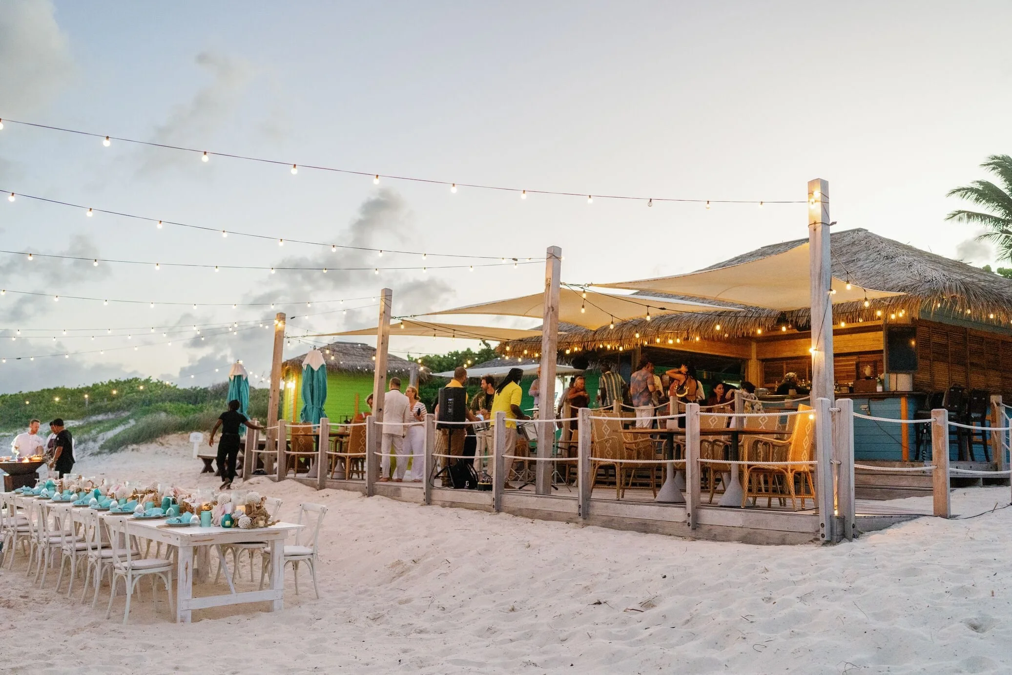 Beachside party scene during sunset with string lights, people socializing, and a thatched roof bar area.
