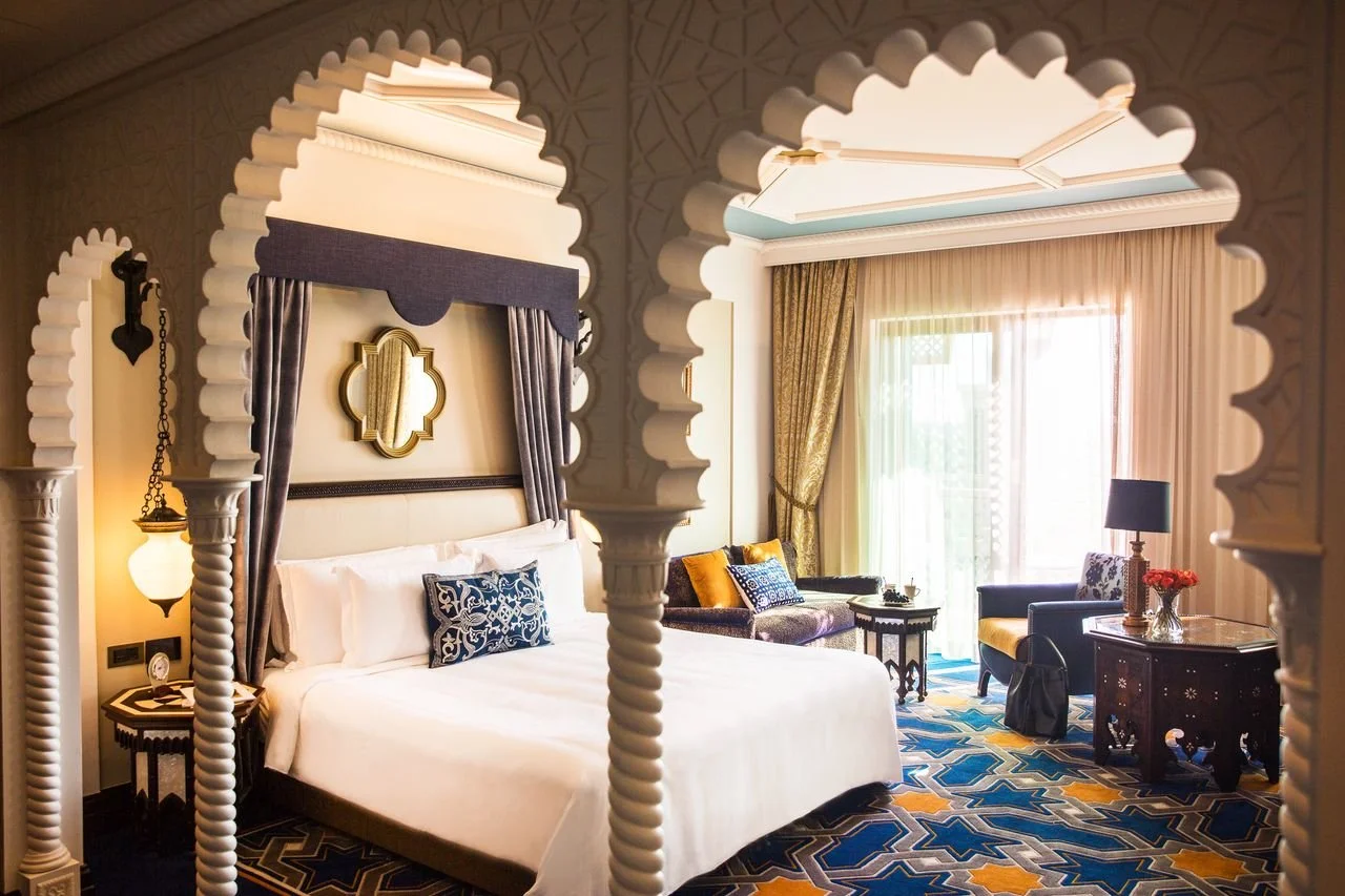 Luxury hotel room with a four-poster bed, traditional Moroccan-style arches, gold accents, and a sitting area with a couch, armchair, and large window with curtains.