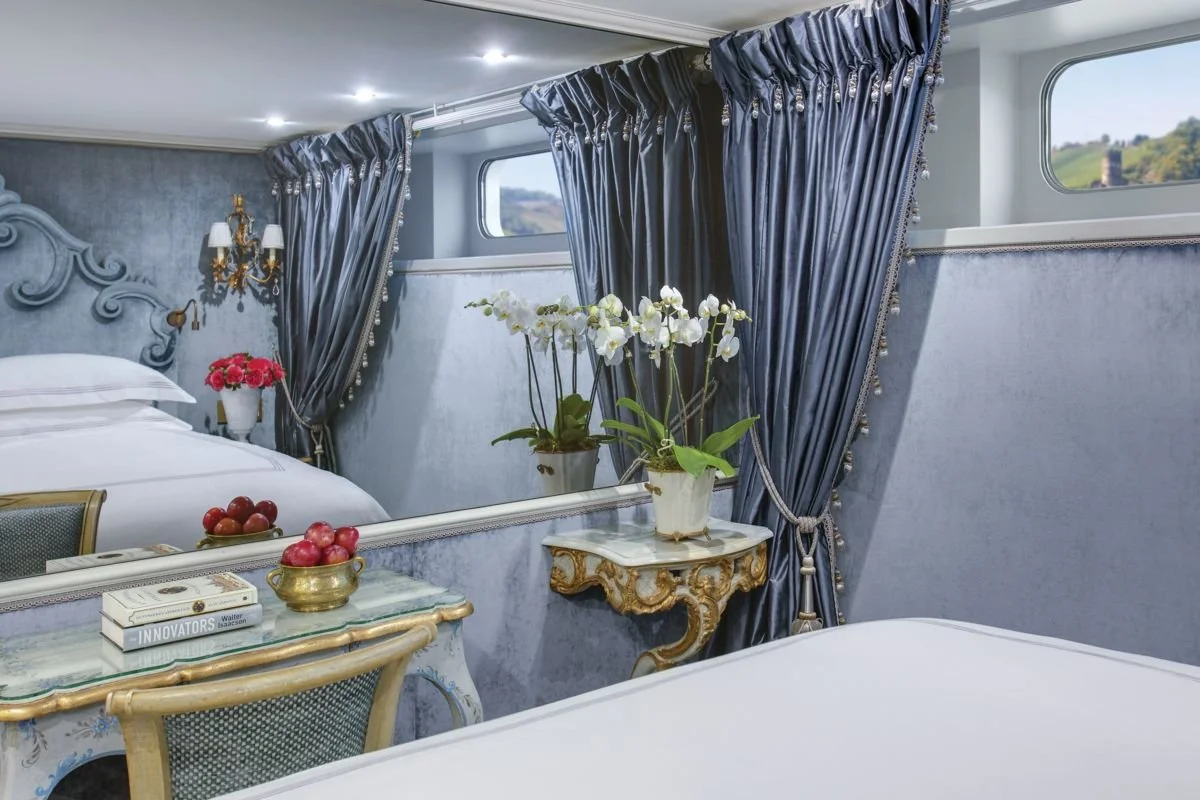 Elegant hotel room with a bed, blue curtains, a mirror, and floral arrangements.