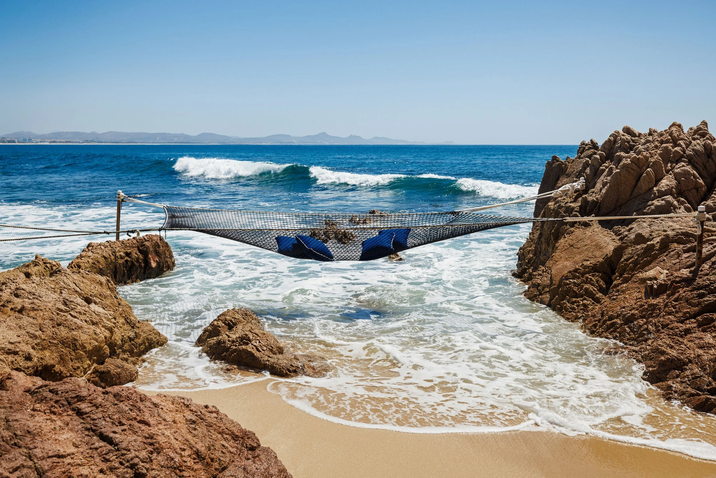 View of rocky beach with water, waves, and a fishing net strung between rocks, with distant mountains on the horizon under a clear blue sky.