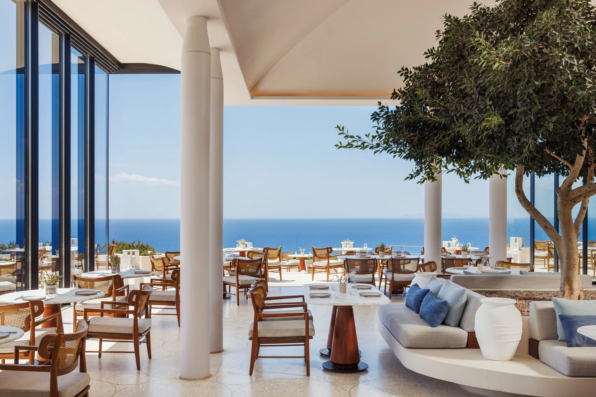 Interior view of a modern seaside restaurant with white walls, large glass windows, wooden chairs, white tables, and a view of the ocean and sky outside.