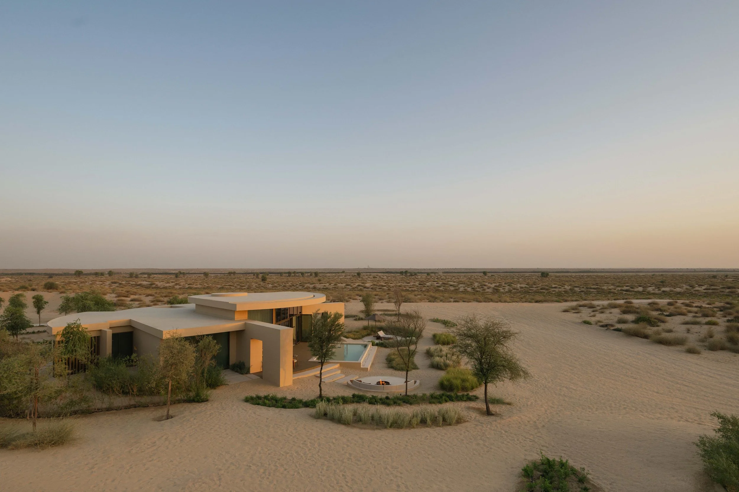 Modern house with flat roof and large glass doors, situated in a desert landscape with sparse trees and bushes, under a clear sky.