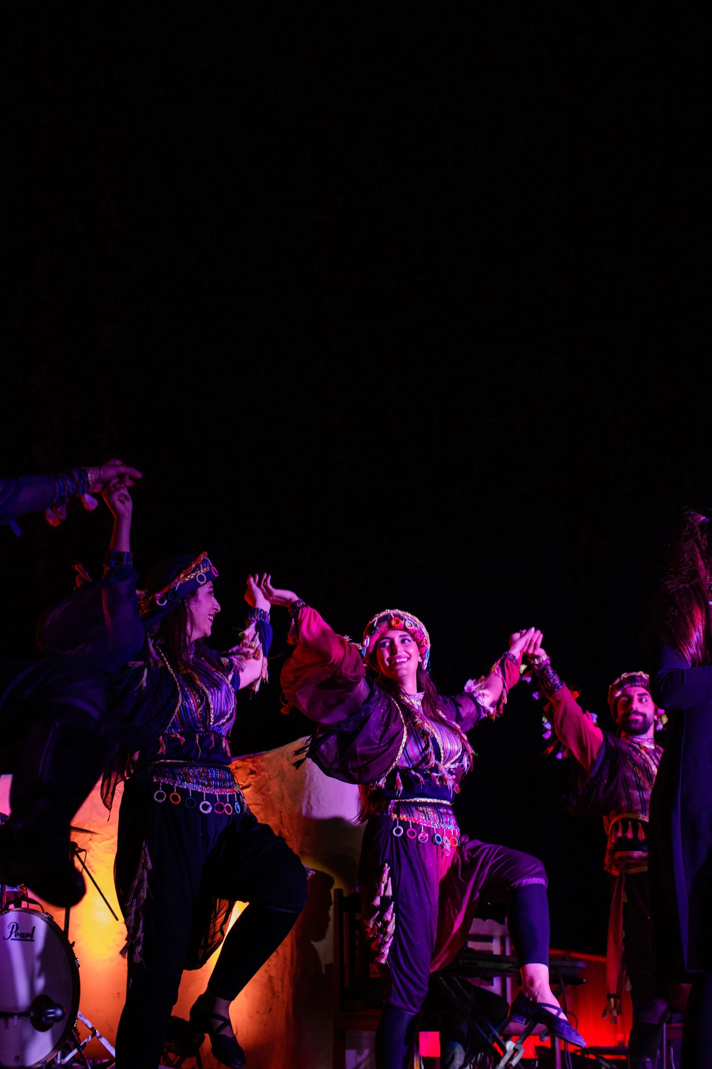 Group of performers dressed in traditional costumes dancing on stage under colorful lighting, with some musical instruments visible.