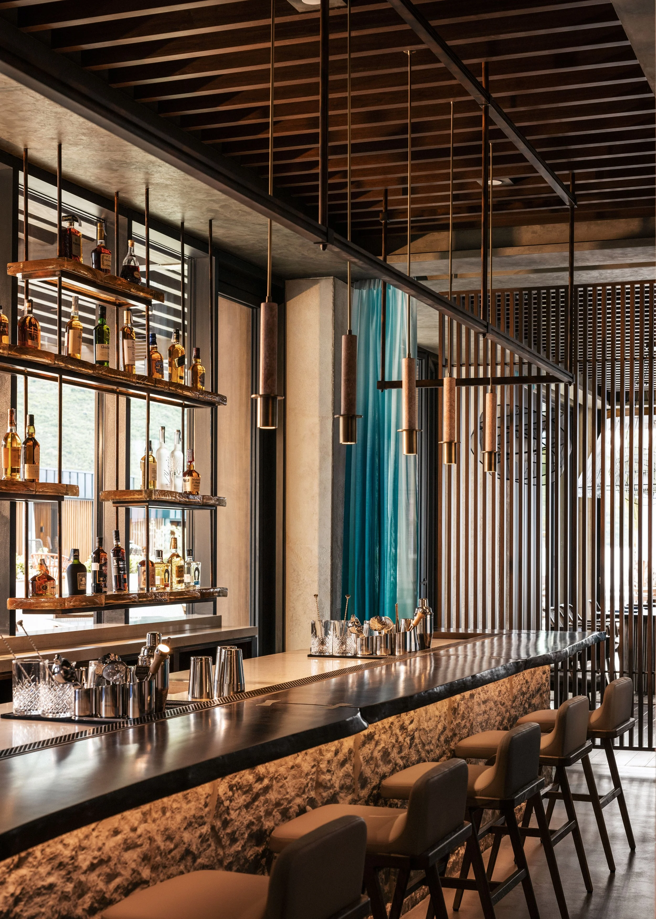 A modern bar with a marble countertop, bar stools, and shelves of liquor bottles; hanging pendant lights and wooden ceiling design.