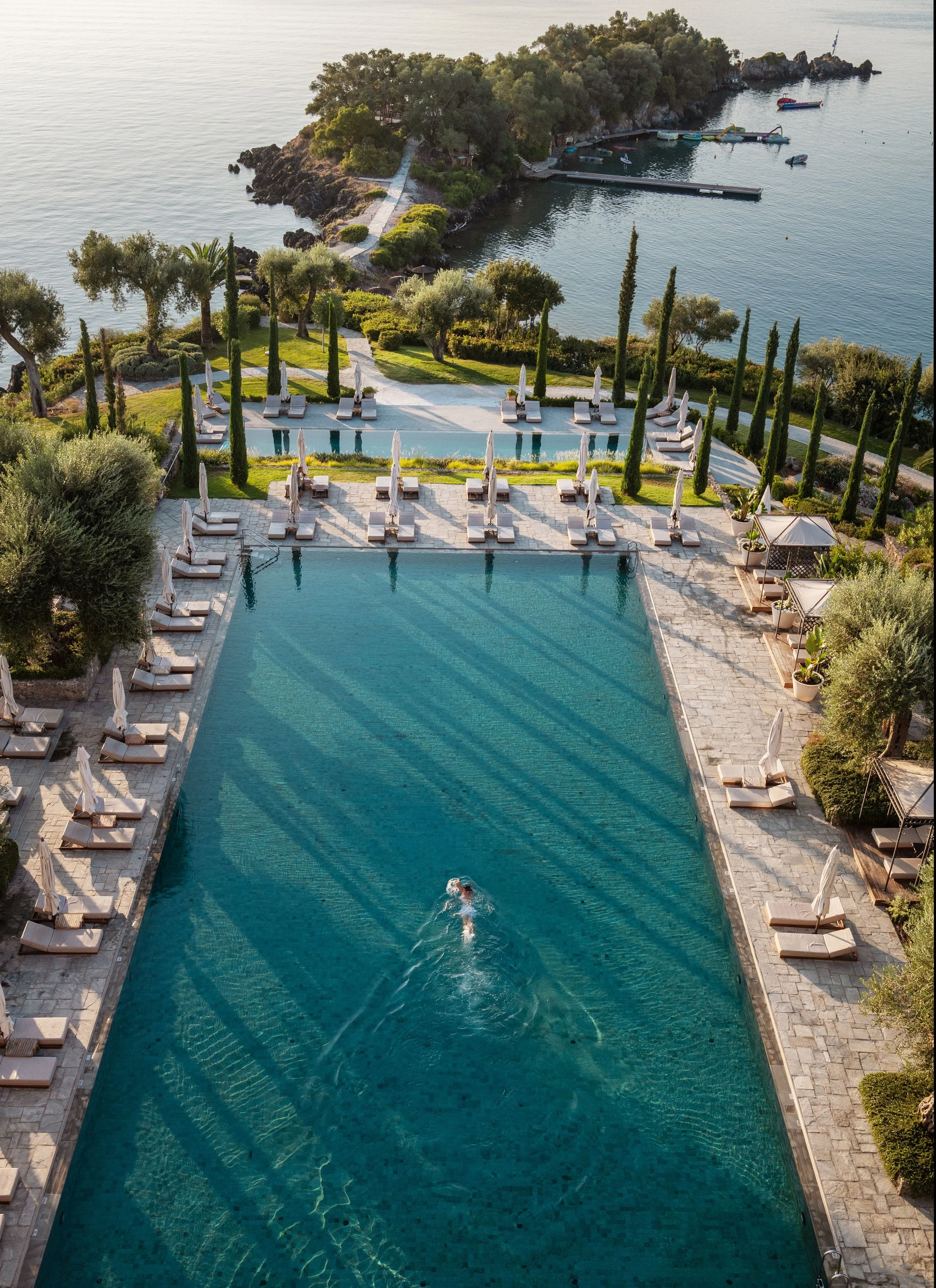 Aerial view of a swimming pool area at a seaside resort with lounge chairs, umbrellas, and lush greenery. The pool extends towards the water, with a swimmer in the pool. In the background, there is a coastline with trees, rocky outcrops, and small boats docked near the shoreline.