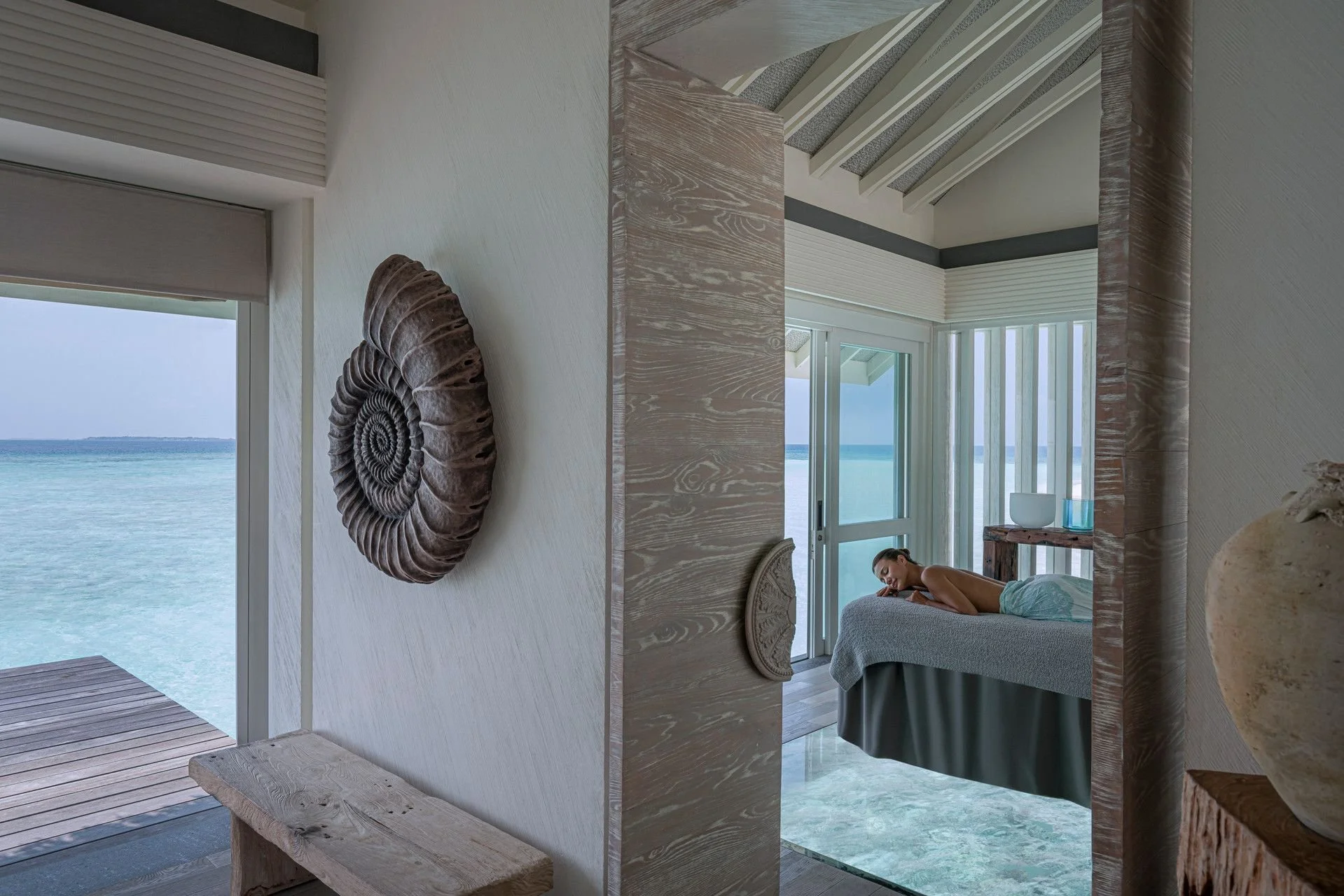 A woman lying on a bed in an oceanfront bedroom with large windows, a wooden bench, and decorative shell artwork on the wall, overlooking the ocean.