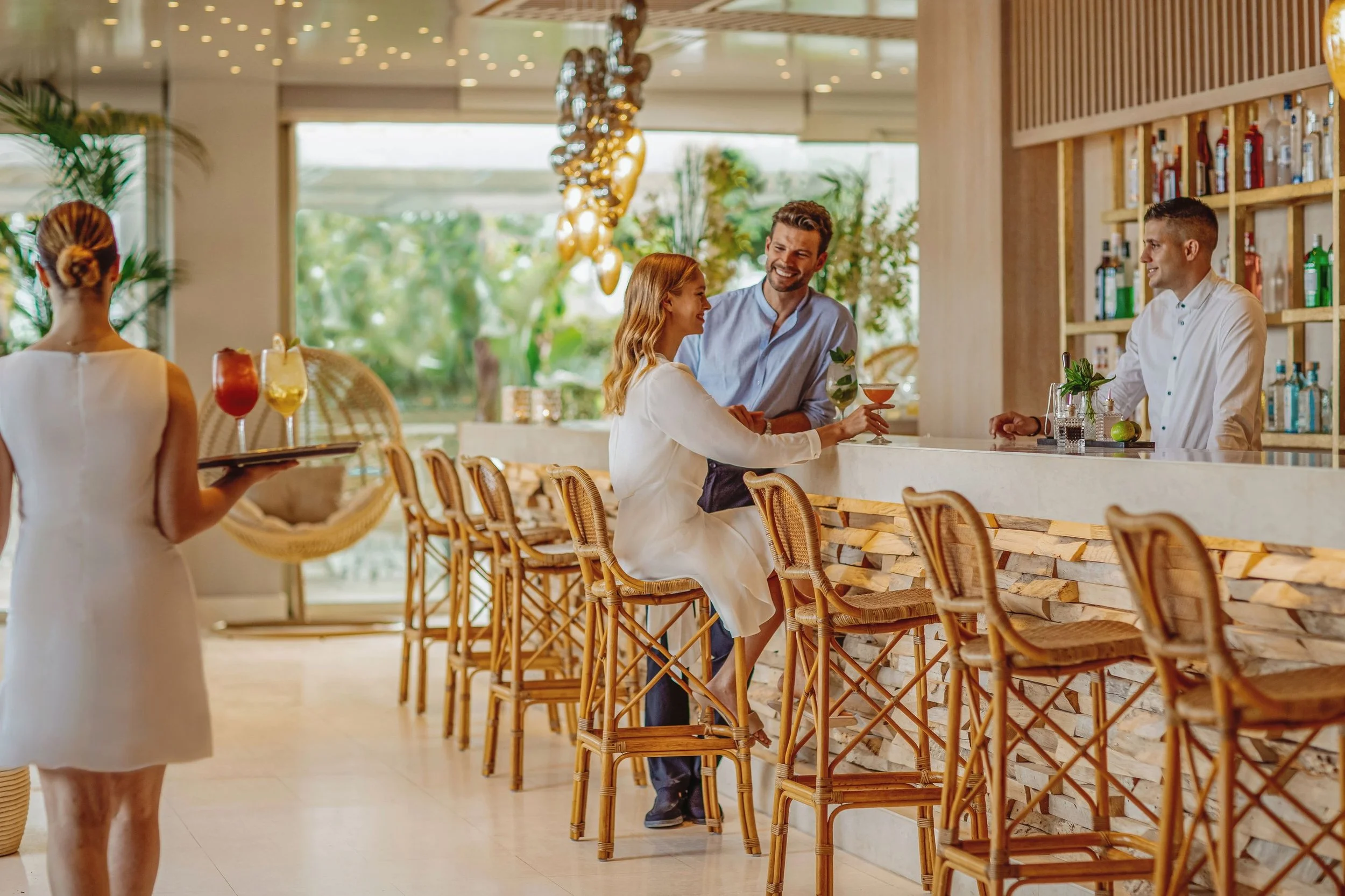 A woman with a bun hairstyle is serving drinks in a white dress at a modern, tropical-themed bar with a wooden bar counter and rattan chairs. Two bartenders are behind the counter, and a couple is sitting, engaging in conversation and holding cocktails. The background features large windows, greenery, and decorative lighting.