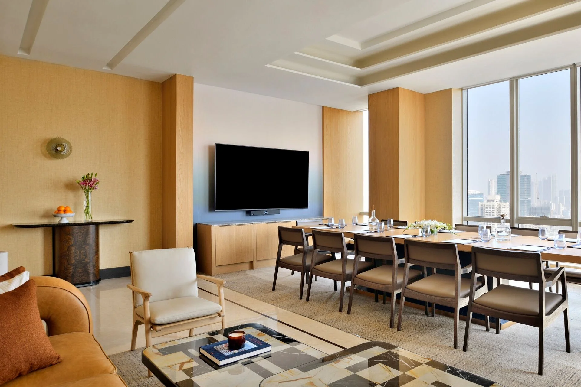 A modern high-rise apartment living and dining room with large windows, a wall-mounted TV, a dining table with chairs, a seating area with armchairs, a coffee table with books and a candle, and city skyline views.
