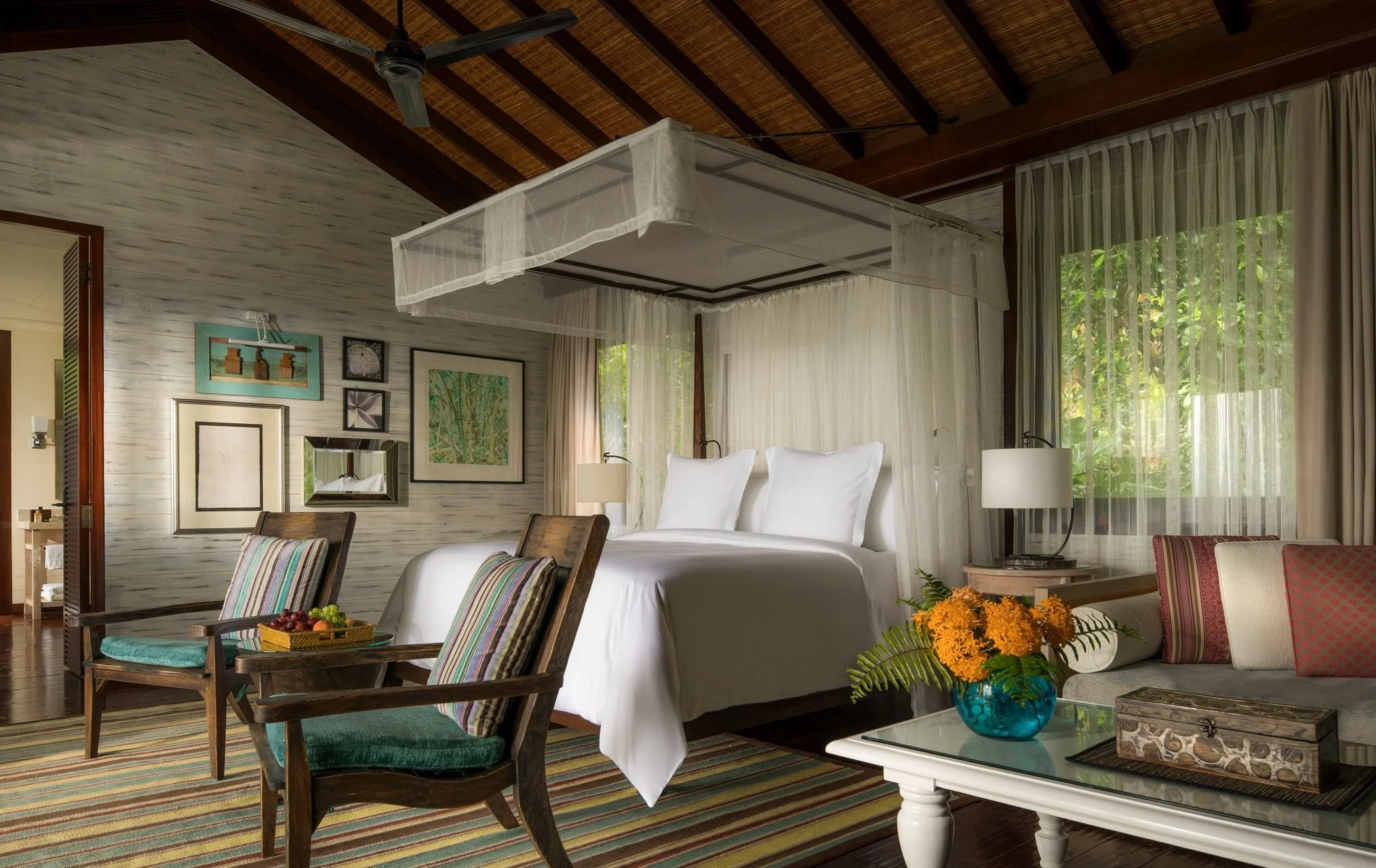 A bedroom with a four-poster bed, white curtains, and framed artwork on a light wood-paneled wall, with windows showing greenery outside.