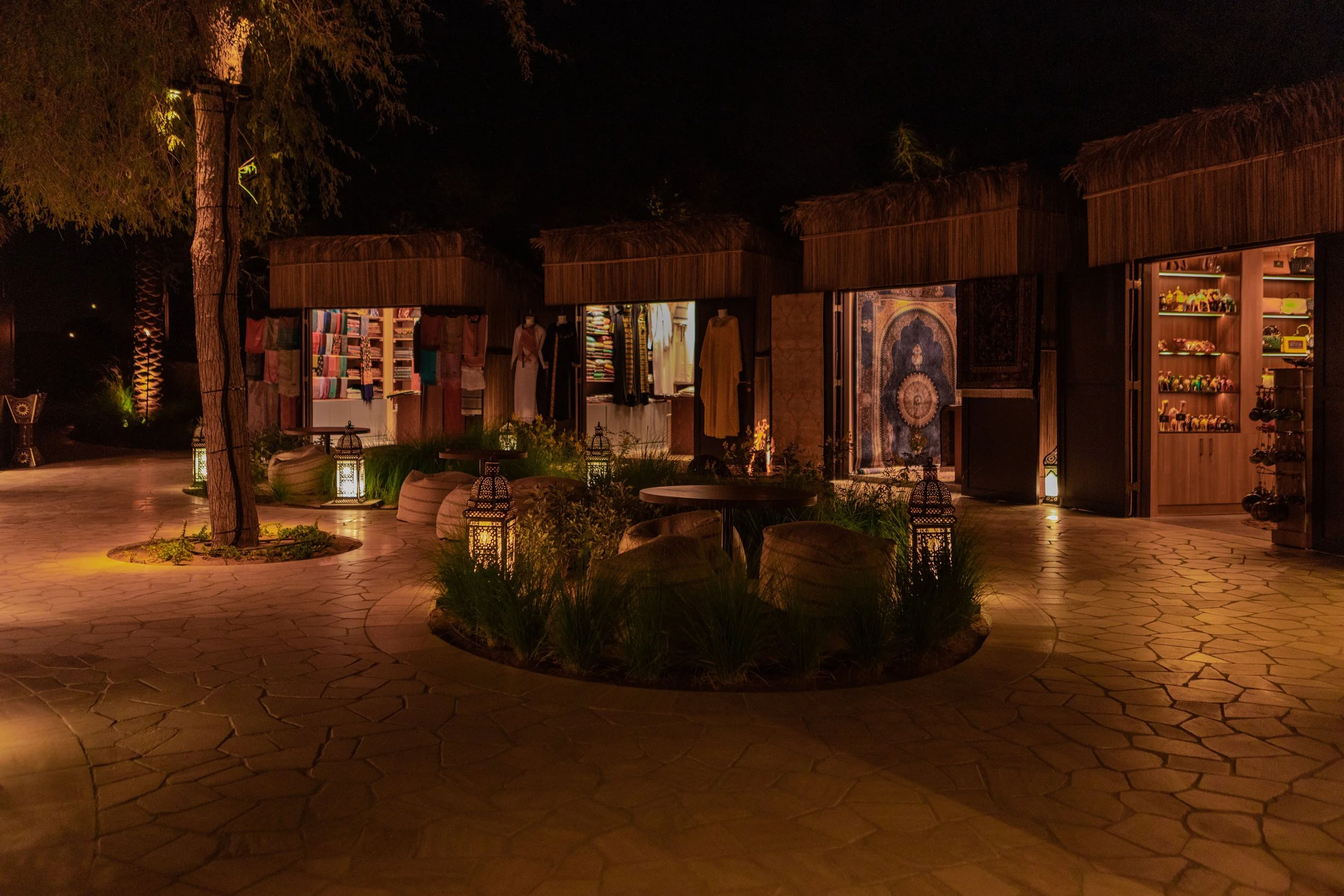 Night view of an outdoor market with small shop stalls made of wood and thatch, lit by warm lanterns, with potted plants and trees around.