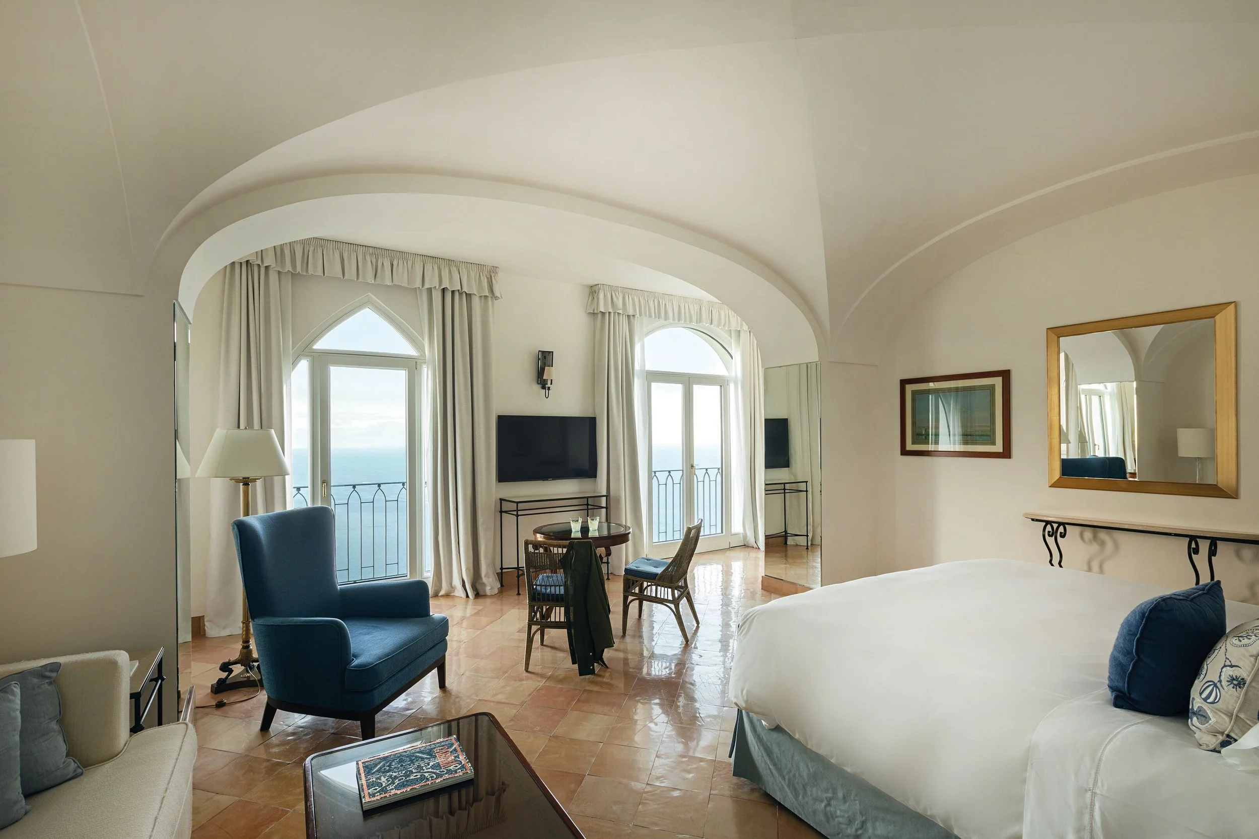 A spacious hotel room with white walls and ceiling, large arched windows with curtains revealing ocean views, and a mix of seating including a blue armchair, sofa, and dining chairs. There is a mirror on the wall, a small table, and a bed partially visible on the right.