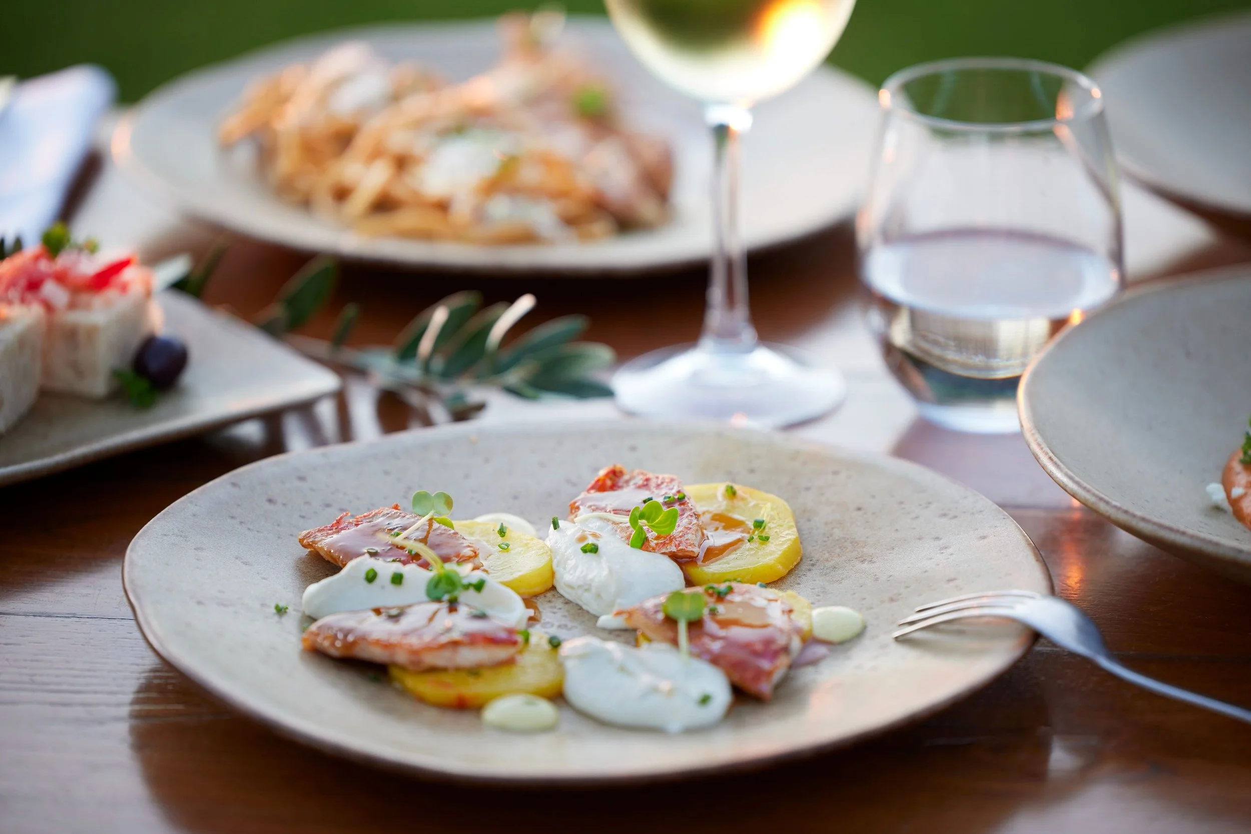 Close-up of a plate with sliced potatoes, poached eggs, ham, and chopped herbs on a wooden table with other plates and glasses of water and wine in the background.
