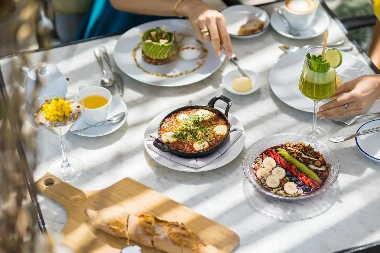A table set with various dishes including a bowl of gazpacho, a berry smoothie bowl, an avocado toast, a fruit salad with bananas and berries, a green cocktail garnished with mint and lime, a cup of tea, and a small dish of butter. A person's hand is reaching for the cocktail while another hand is serving itself from a skillet of shakshuka.
