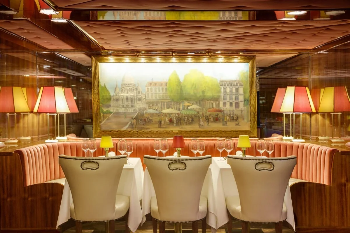Elegant restaurant with a round table set for dinner, surrounded by white chairs with black trim, pink banquette seating, and large pink and yellow lamps. A large framed painting of a city scene with trees and outdoor market is on the wall behind the table.