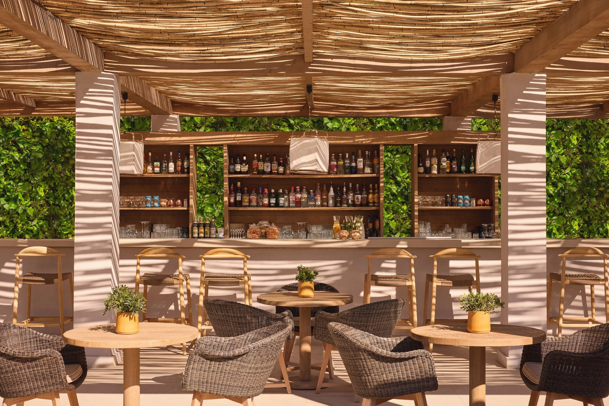 An outdoor bar area with a thatched wooden roof, various bottles of alcohol on shelves, and wicker chairs and small tables with potted plants in front.