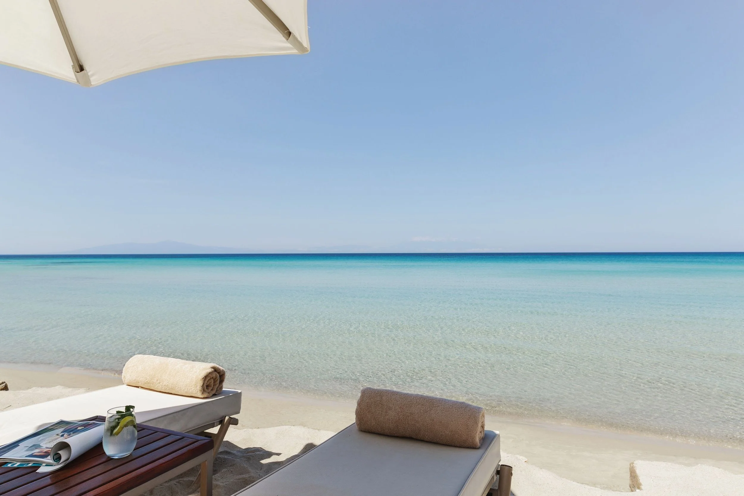 Beach scene with lounge chairs under an umbrella overlooking clear blue ocean and sky, with a rolled towel on a lounge chair and a glass of water with lemon and mint on a small table.
