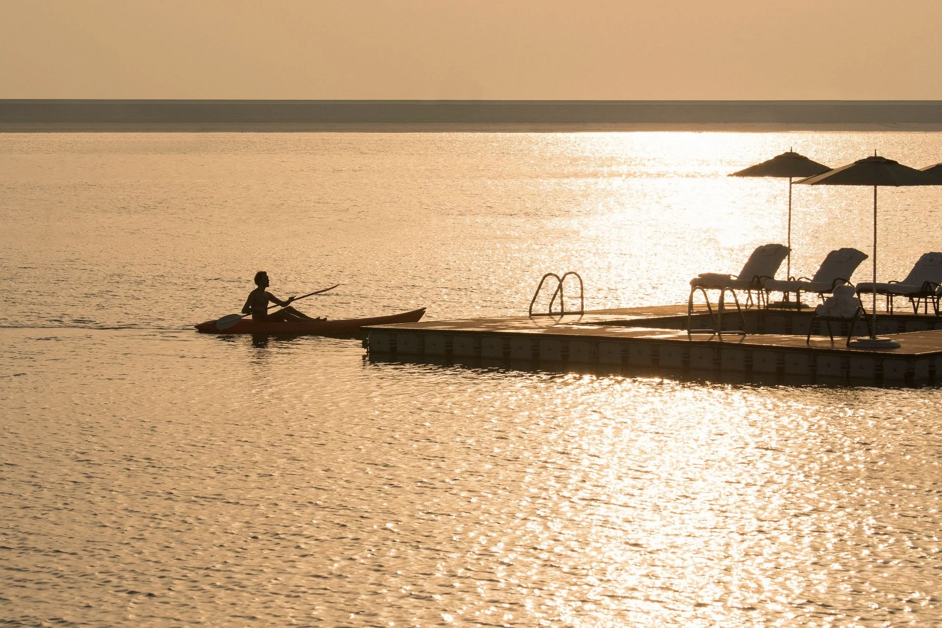 A person paddling a kayak on a calm body of water during sunset with a dock, chairs, and umbrellas on the right and a distant shoreline in the background.