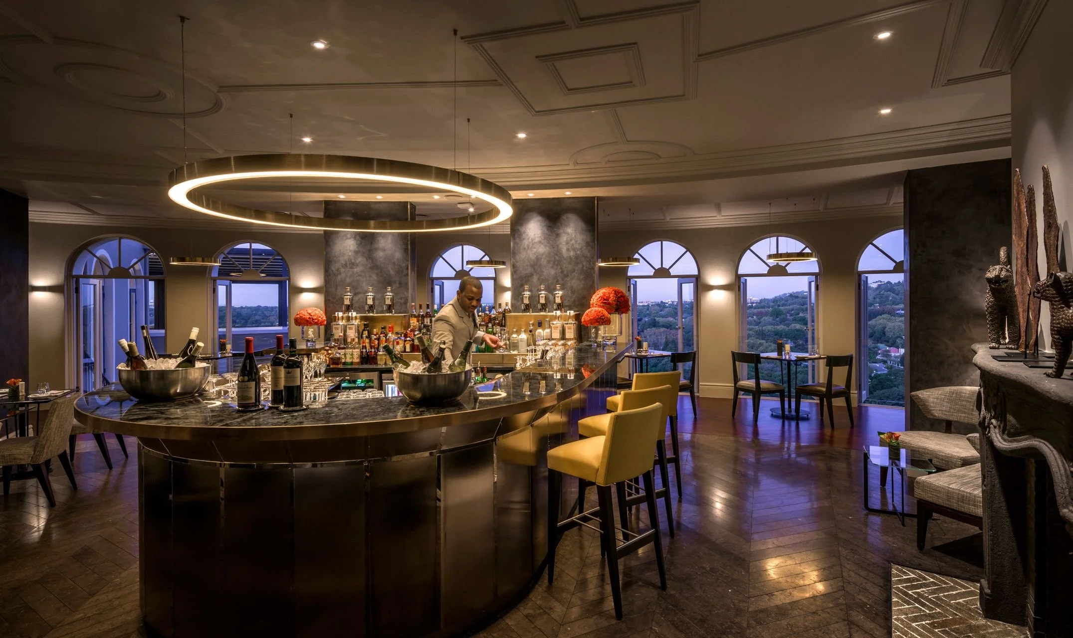 Elegant indoor bar with a curved marble counter, modern circular light fixture overhead, large windows with cityscape view, decorated with red flowers, and a bartender preparing drinks.