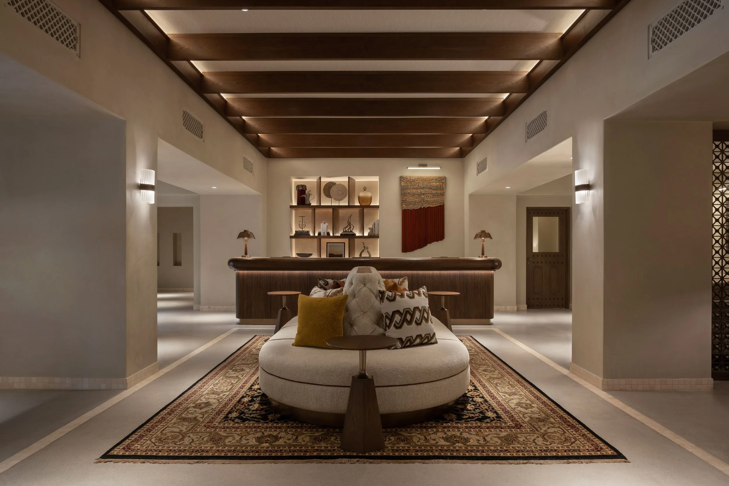 A hotel lobby with modern decor featuring a central cushioned seating area, decorative pillows, wooden accents, a rug, lamps, and artwork on the wall.